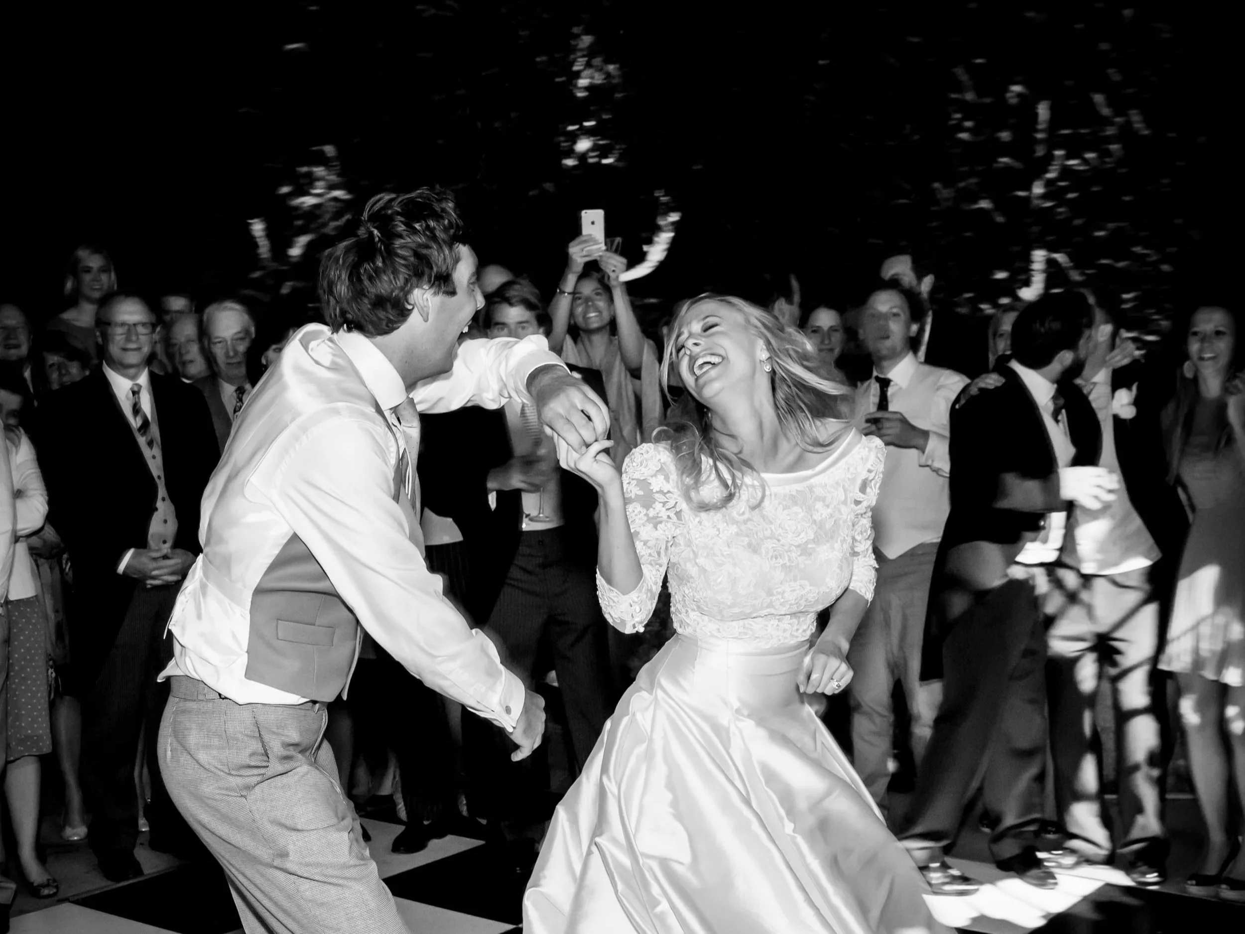 A bride and groom dancing and laughing at their wedding reception, surrounded by guests.