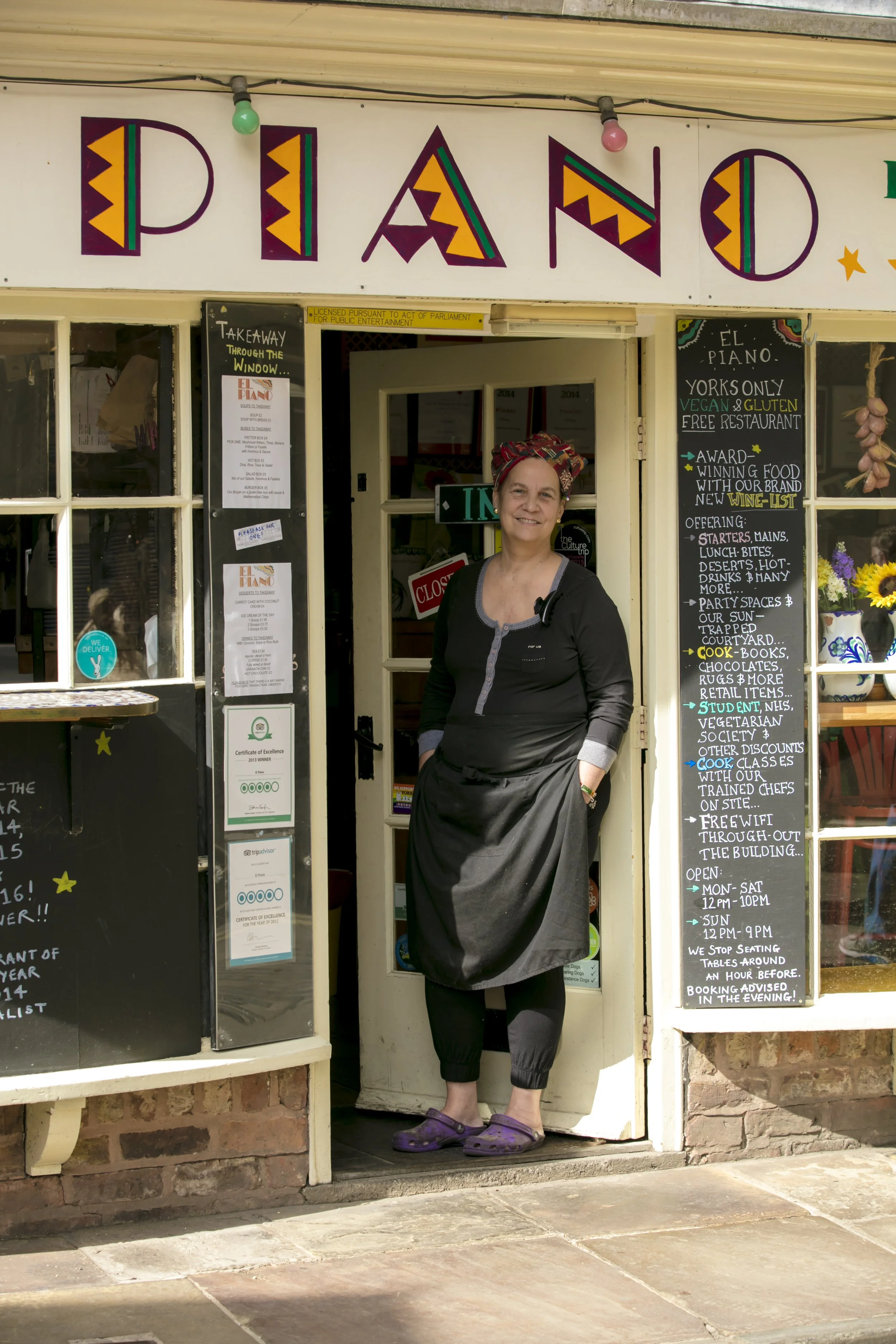 A woman standing in the doorway of a small restaurant or café, smiling at the camera. The establishment has a colorful sign with the word 'Piano' at the top, and menus and notices displayed around the entrance.