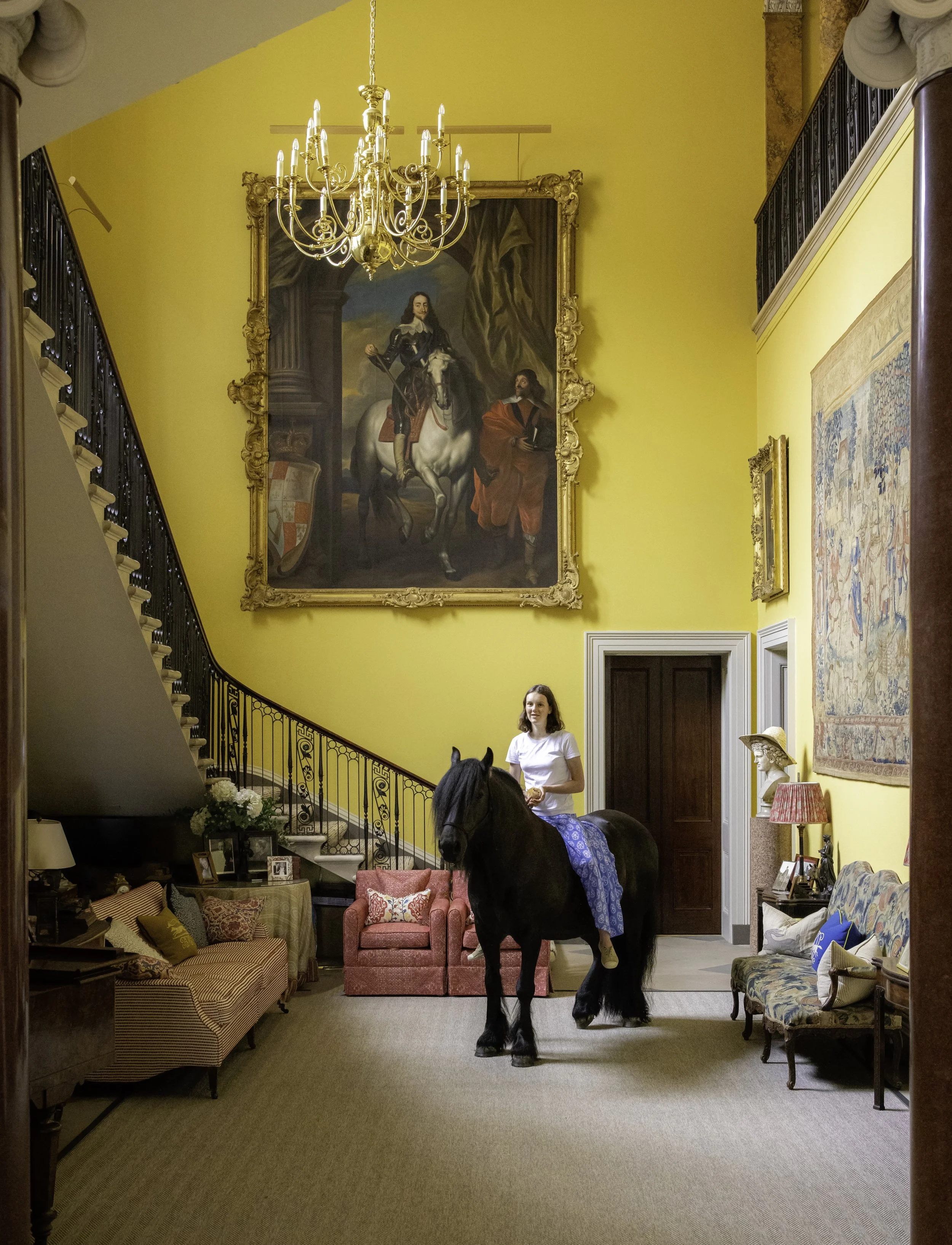 A woman in a white shirt and blue skirt sits on a black horse inside a living room with bright yellow walls, ornate chandelier, and large framed painting of a man on a horse on the wall.