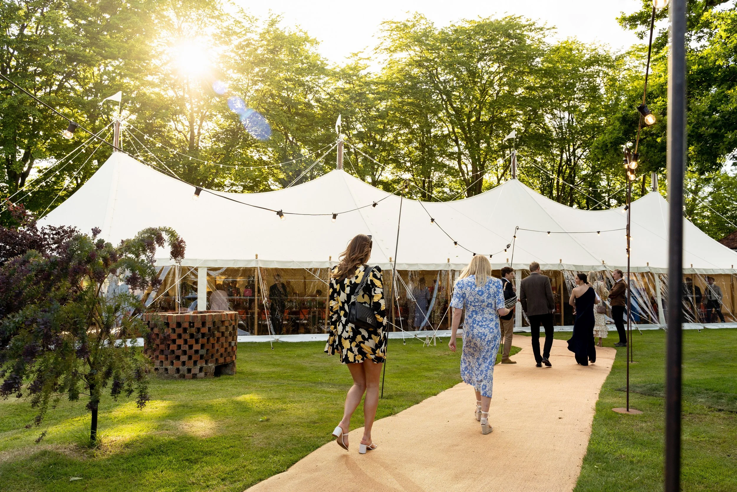 Outdoor event with a large white tent, string lights, and people walking on a beige path on a sunny day with green trees in the background.