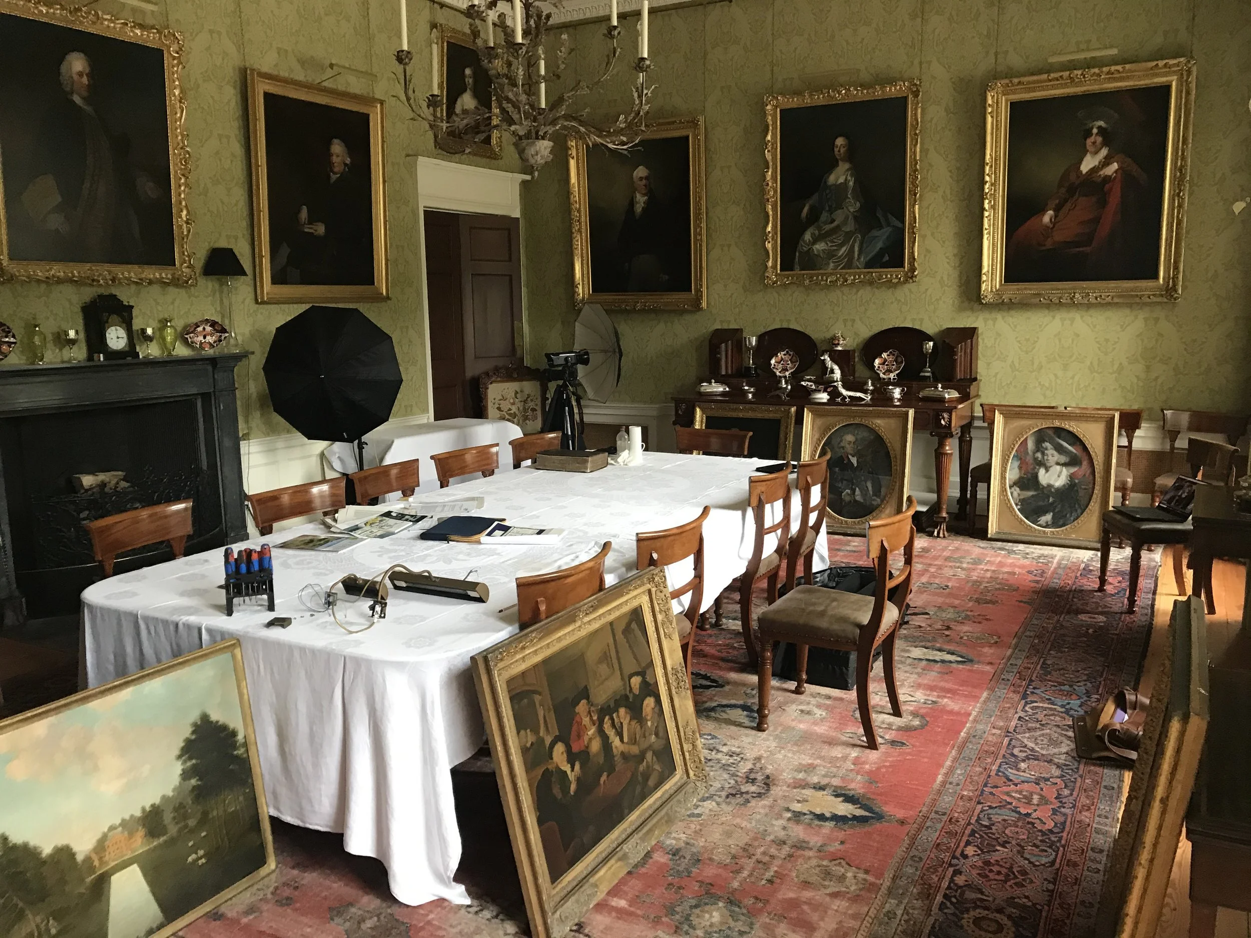 Vintage photo gallery room with large framed portraits on the wall, paintings leaning against the table, a big chandelier, and various antique items.