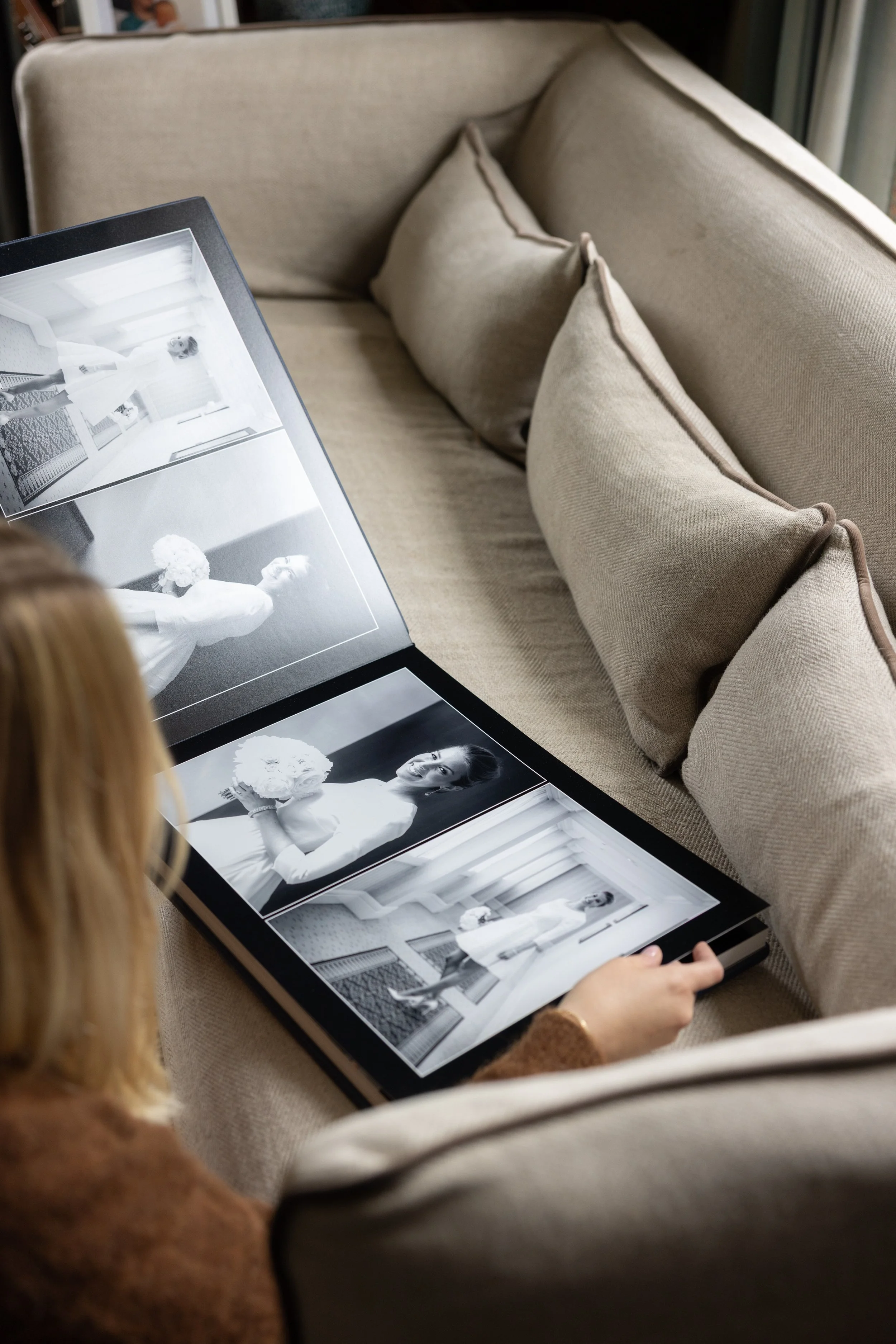 A woman looks at a photo album with black-and-white wedding photos, sitting on a beige sofa with matching cushions in a well-lit room.
