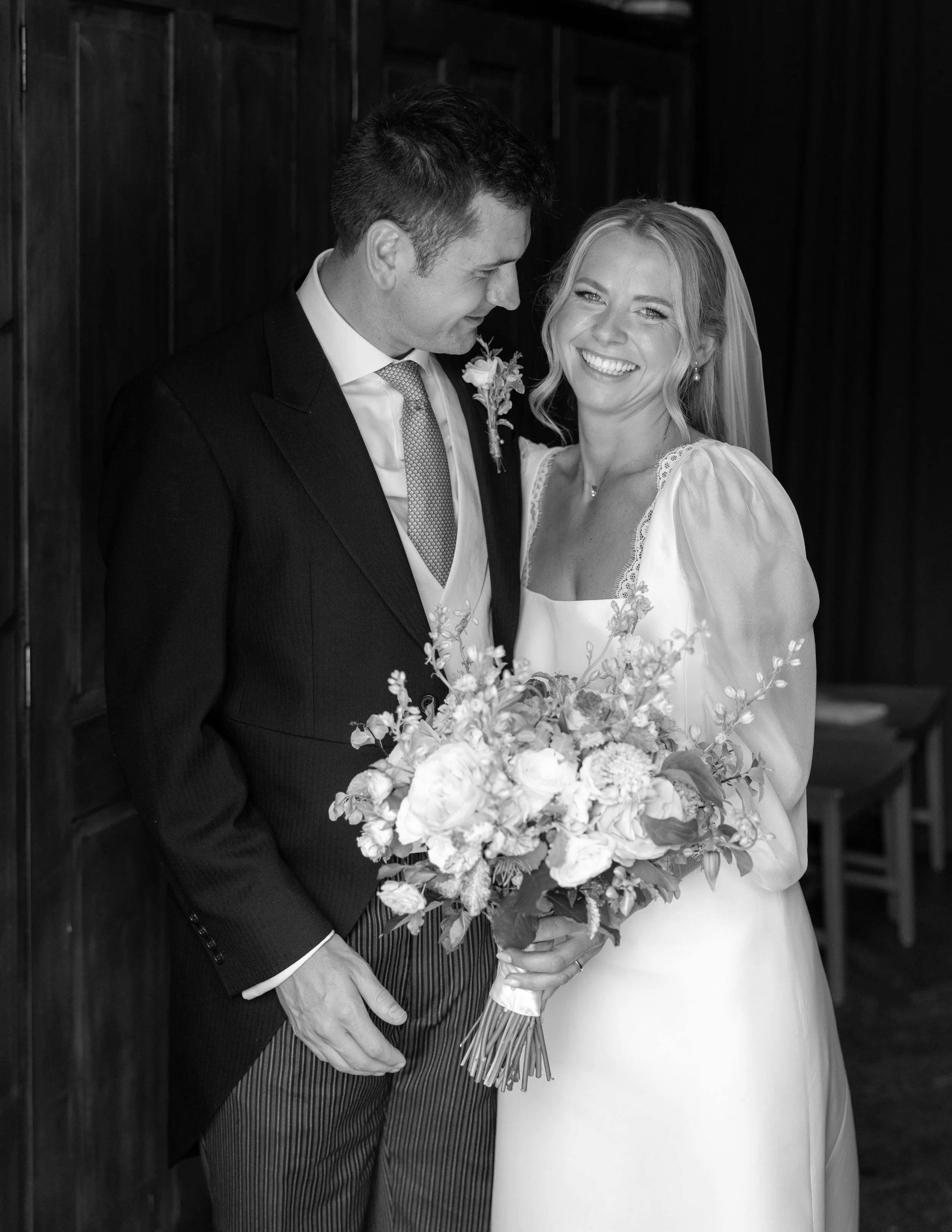 Black and white photo of a bride and groom smiling and looking at each other, with the bride holding a large bouquet of flowers.