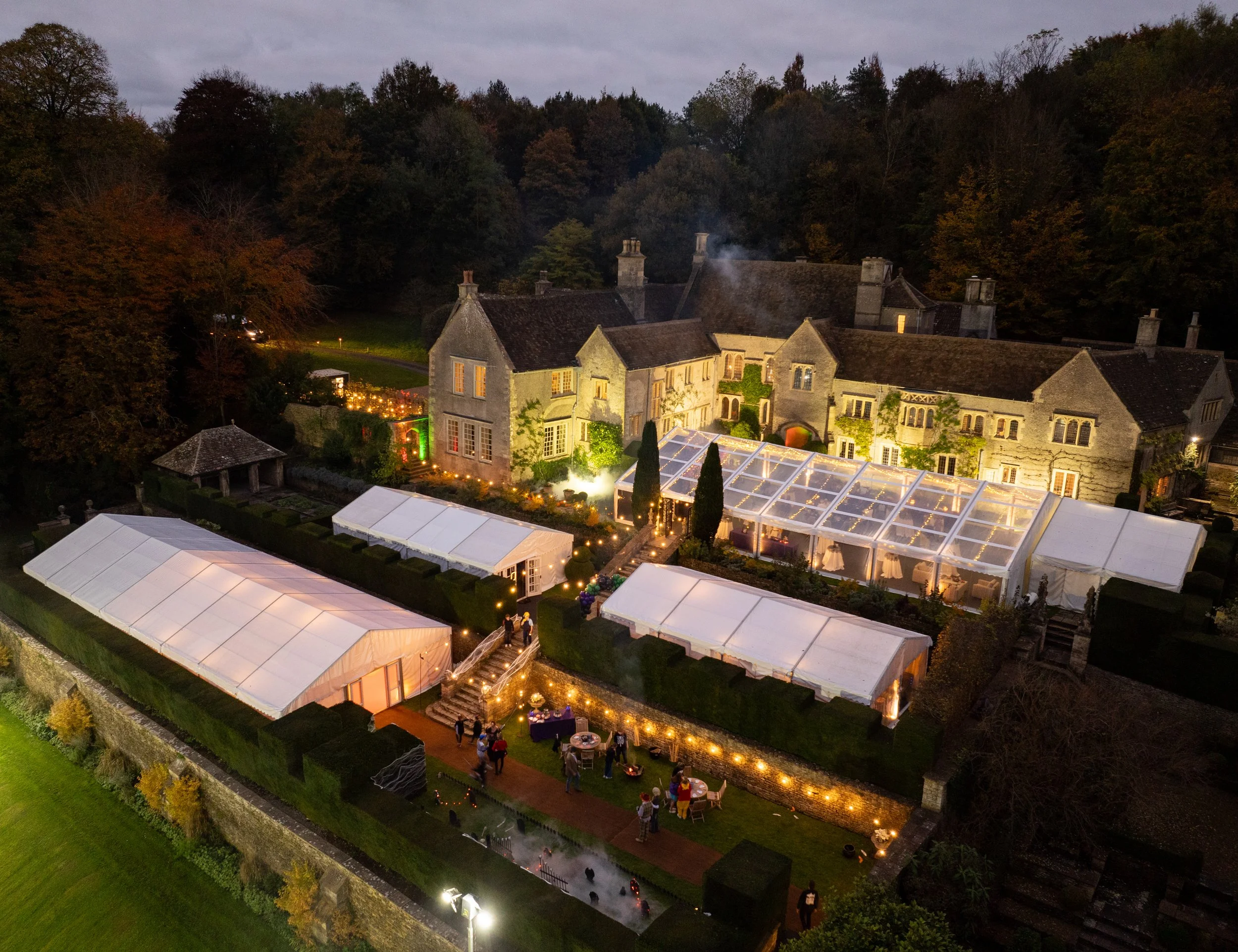 Aerial view of a large mansion with illuminated outdoor event tents and string lights at dusk, surrounded by trees and greenery.