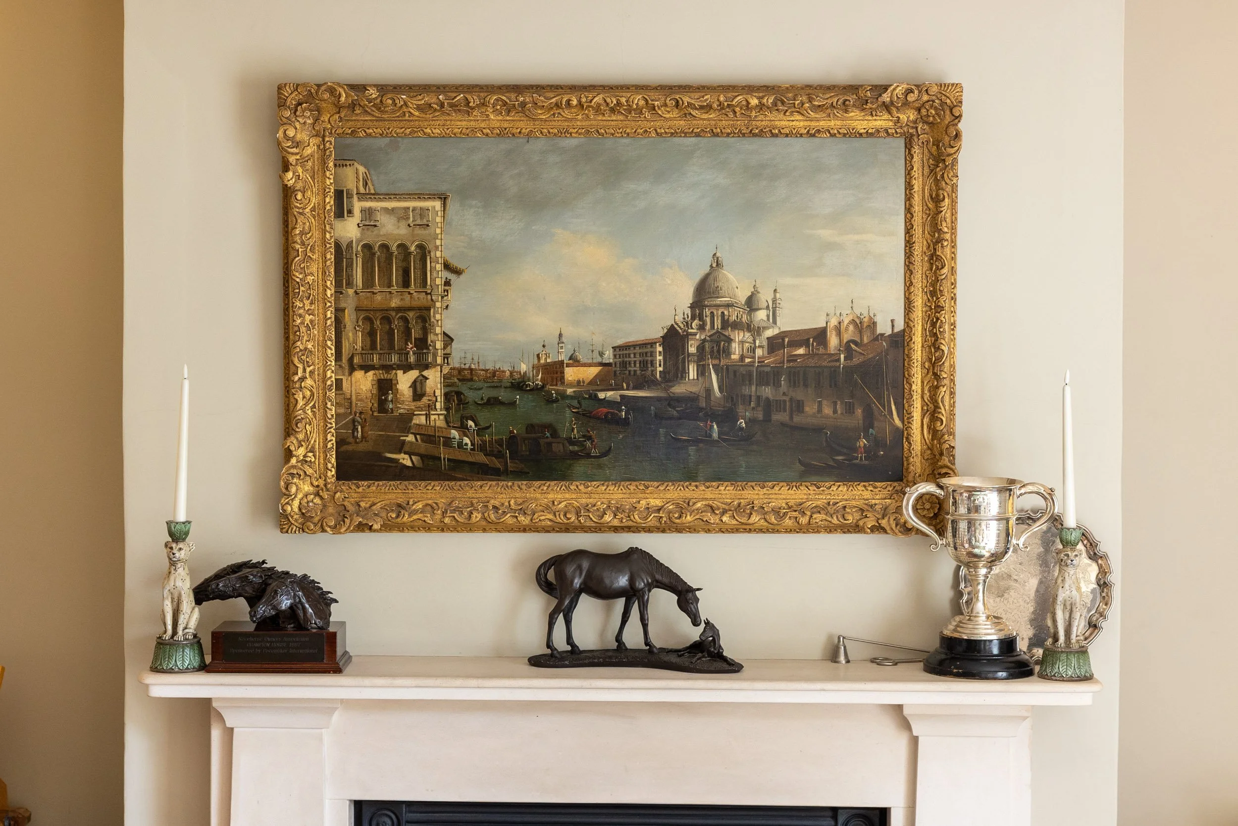 Framed painting of Venice with buildings along a canal, boats, and a cloudy sky hangs above a fireplace. Decor items include candlesticks, a statue of a dog, a horse sculpture, and a trophy cup.
