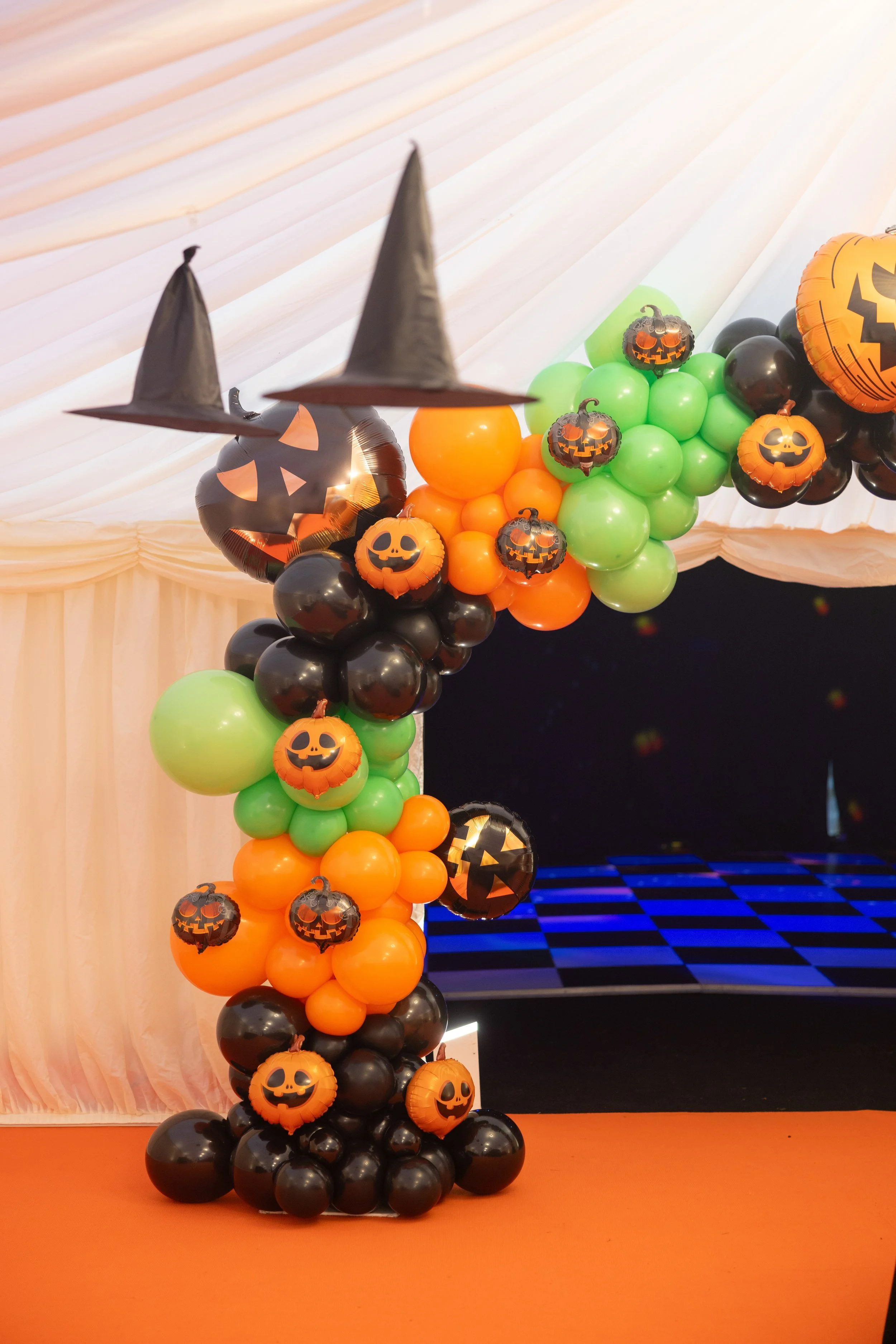 Halloween-themed balloon arch with black, orange, green balloons, pumpkin faces, and witch hats hanging from the top.