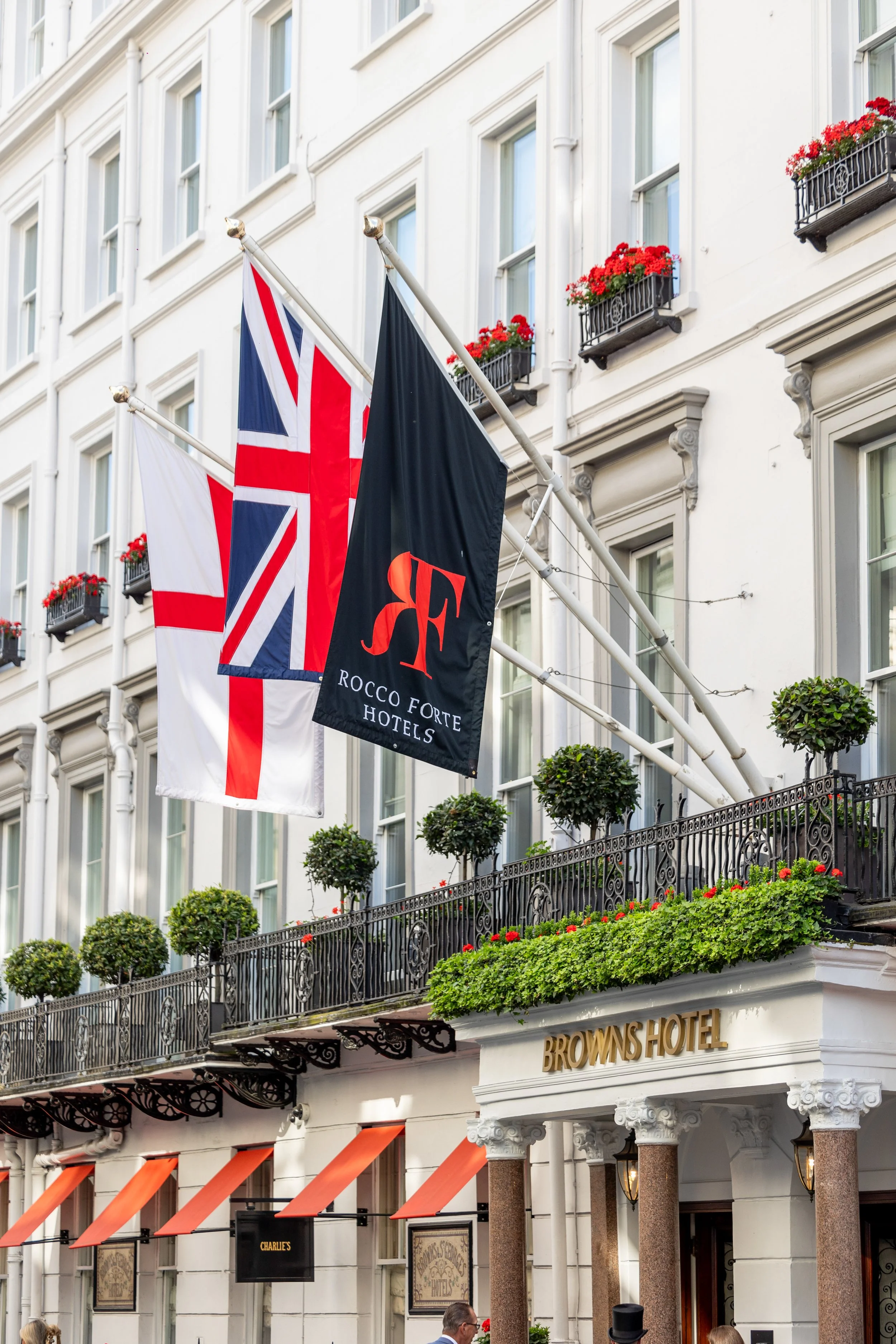 Exterior of the Brown's Hotel with Union Jack, another flag, and a flag with the Rocco Forte Hotels logo flying above a balcony decorated with potted plants and flowers.