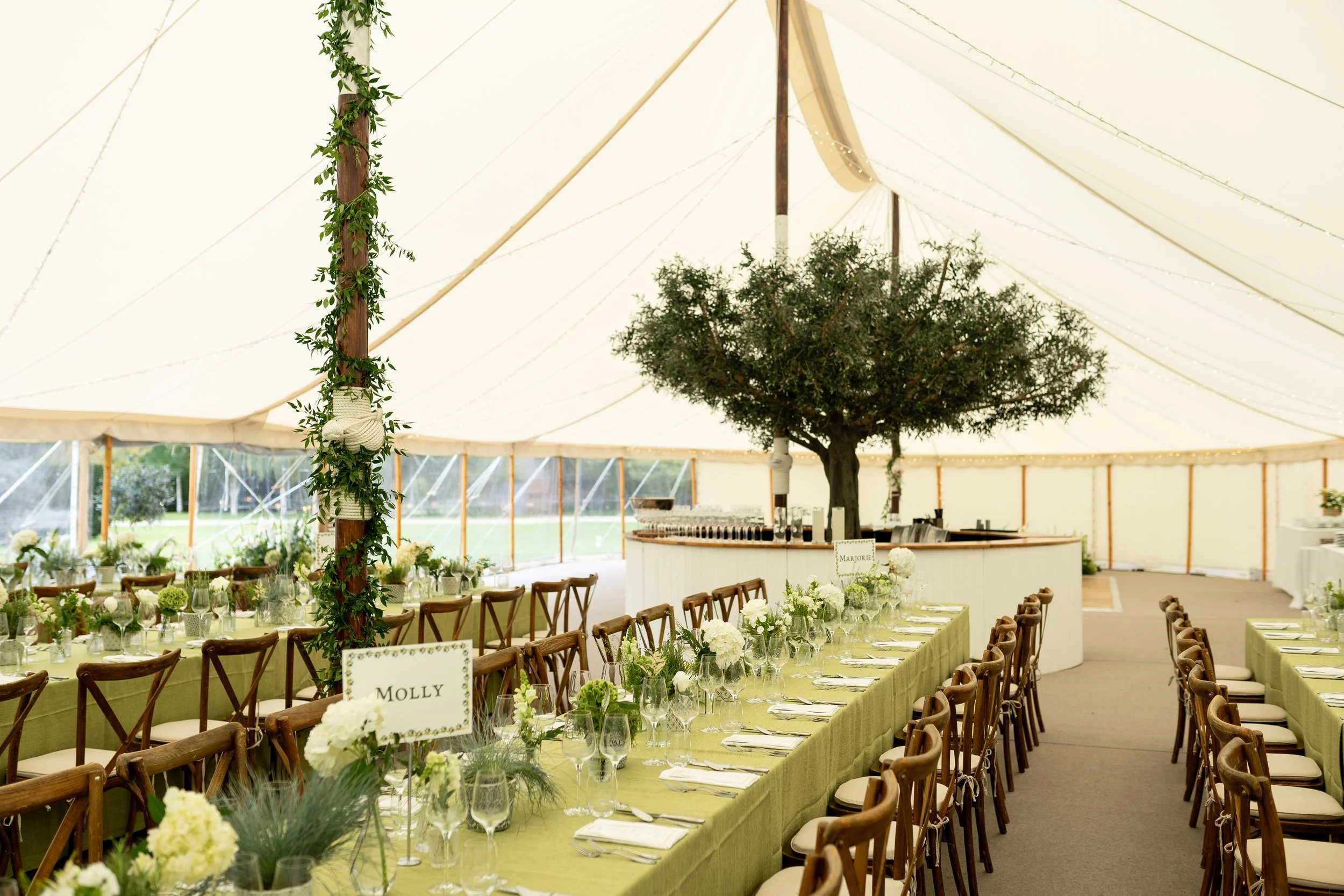 Wedding reception setup inside a large white tent with rectangular tables covered in green tablecloths and decorated with white flowers, napkins, glasses, and silverware. A large tree is centered at the far end of the tent, and there are wooden chair