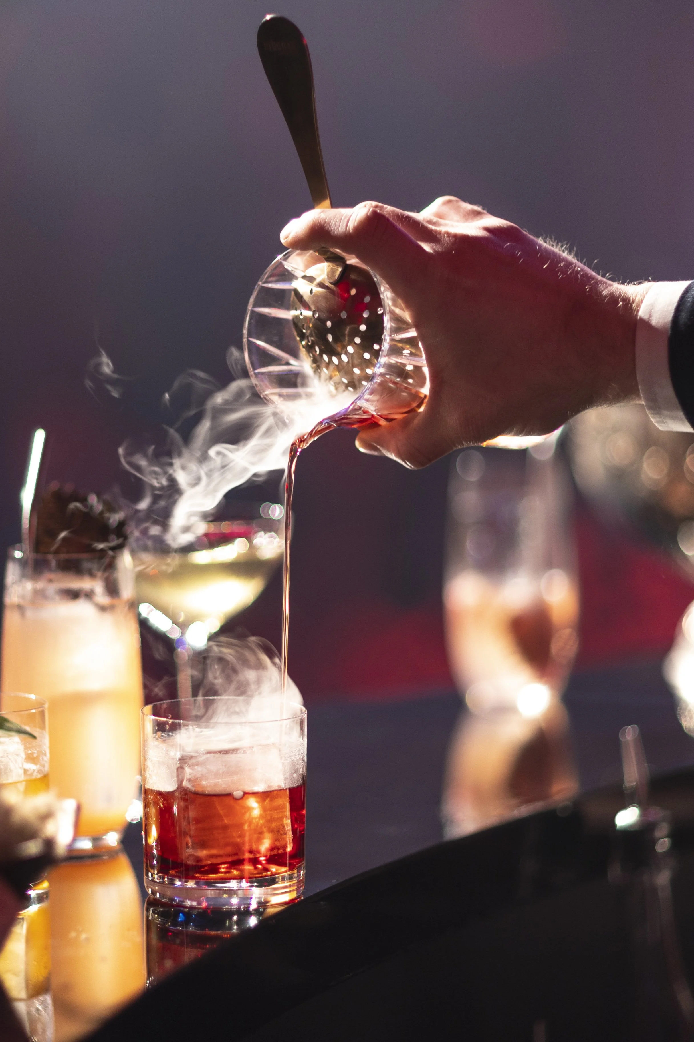 A hand pouring a pink cocktail from a metal shaker into a glass, with other colorful cocktails in the background and steam rising from the drinks.