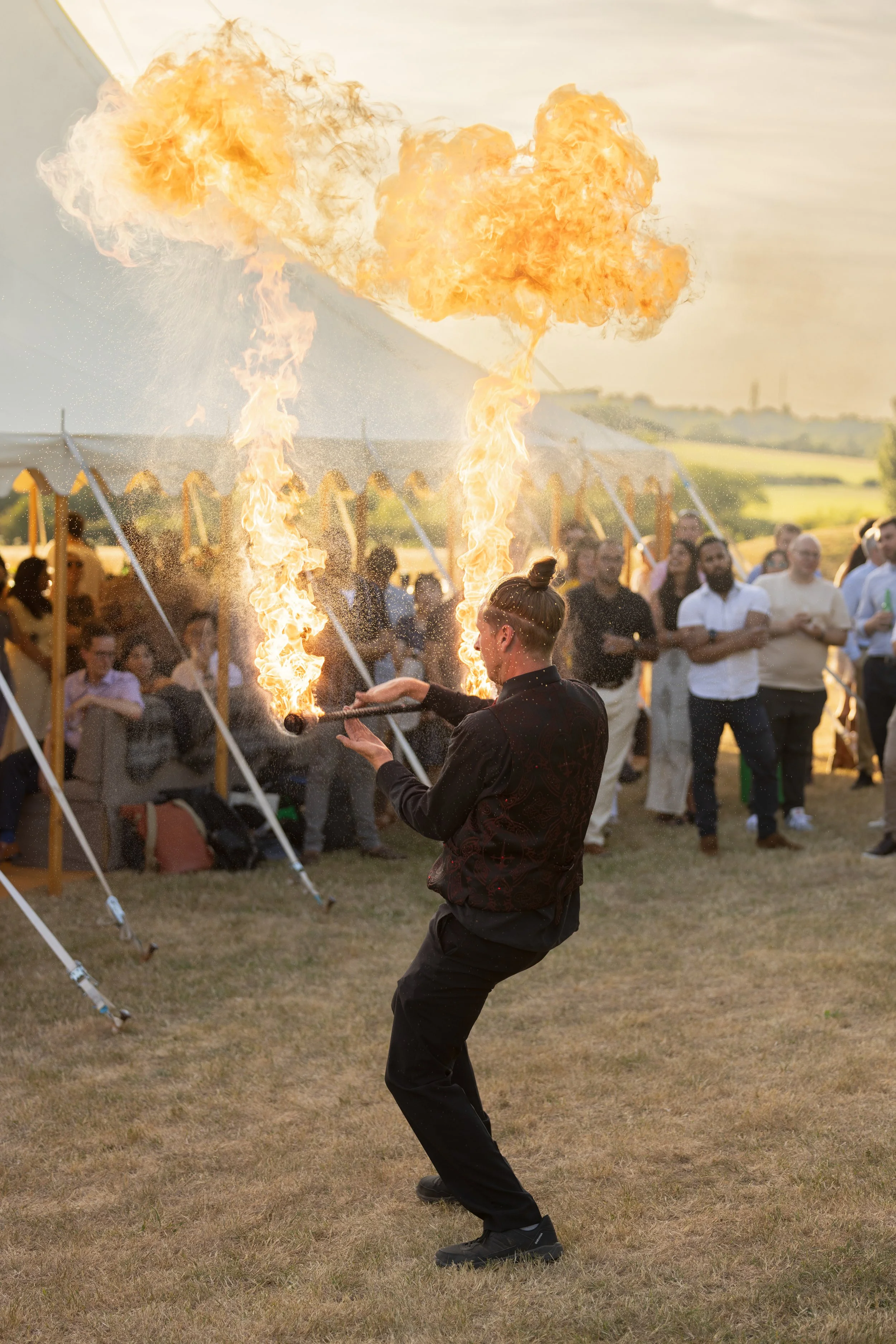 A performer is creating fireballs during a fire show at an outdoor event, with a crowd of onlookers standing behind.