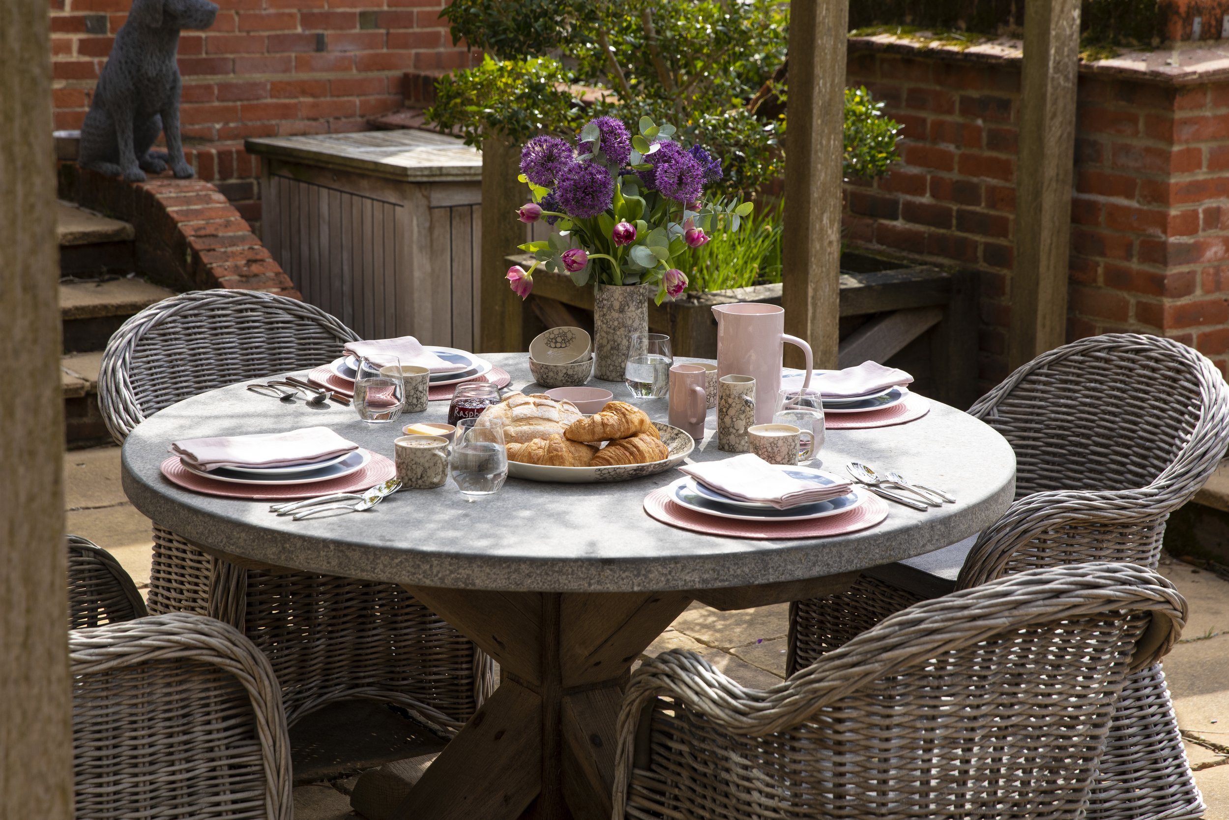 A round outdoor dining table set for breakfast with three plates, cups, glasses, and cutlery, featuring a vase with purple and pink flowers, alongside a plate of croissants and bread, in a garden patio surrounded by brick walls and wooden furniture.