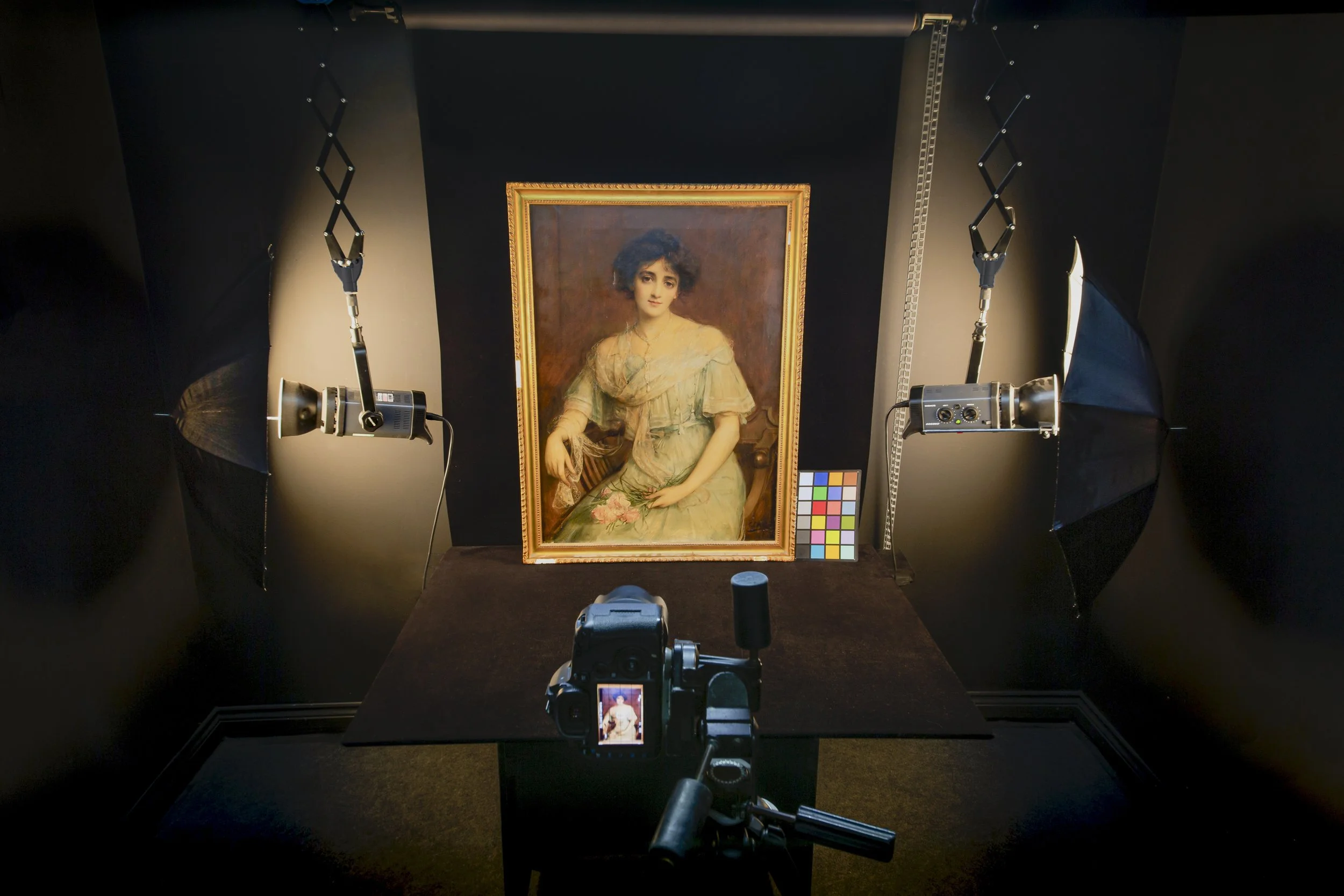 Still life photograph of a studio setup for photographing a framed portrait of a woman. The portrait depicts a woman with dark curly hair in a beige dress. Two softbox lights are positioned on either side of the artwork, and a camera on a tripod is directed at the portrait. A color calibration chart and a white and black scale are visible beside the portrait.