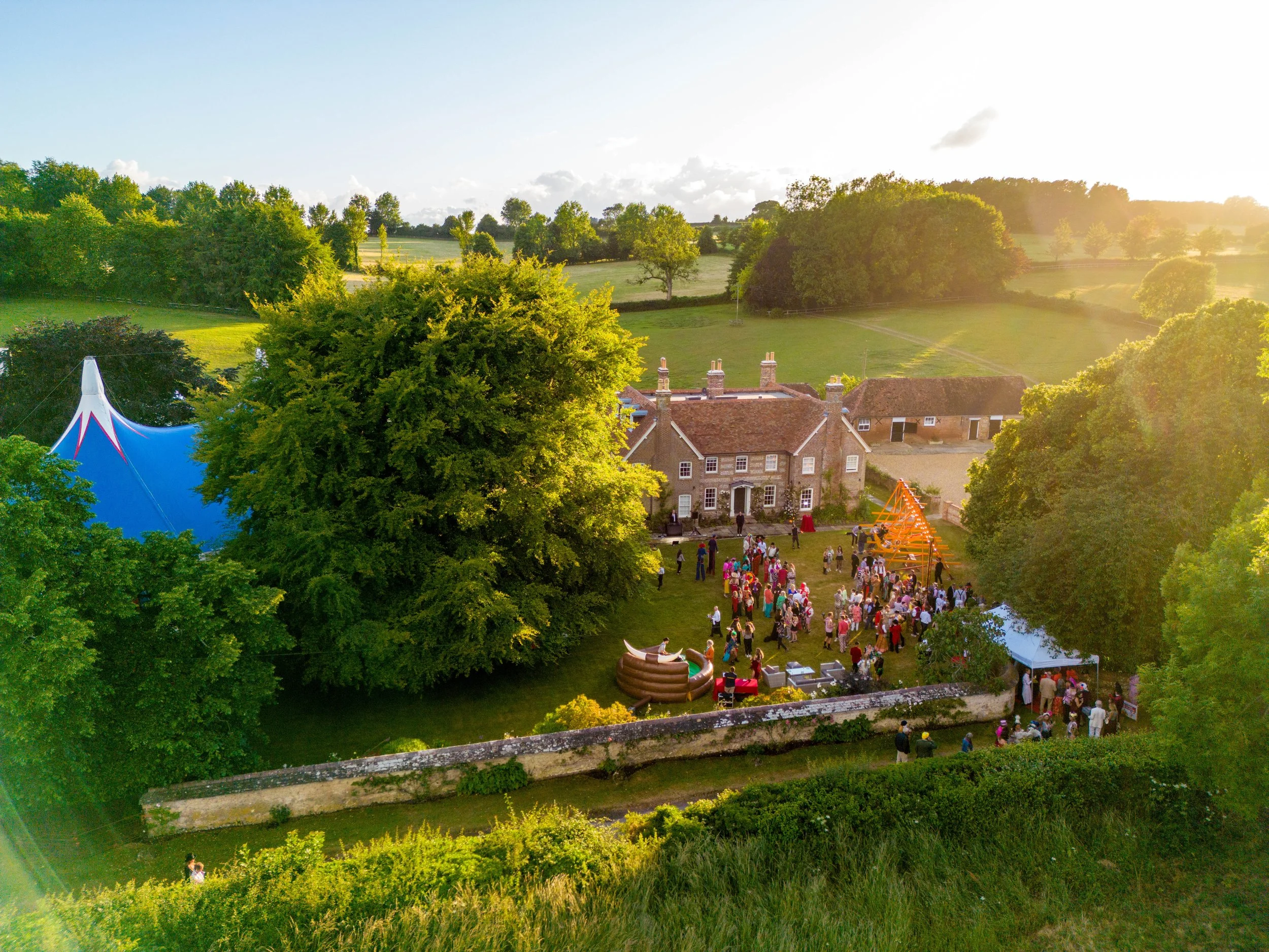 A bird's eye view of an outdoor gathering at a historic mansion during the late afternoon with many people, a large blue circus tent, a small inflatable pool, and orange wooden frames in a lush green garden.