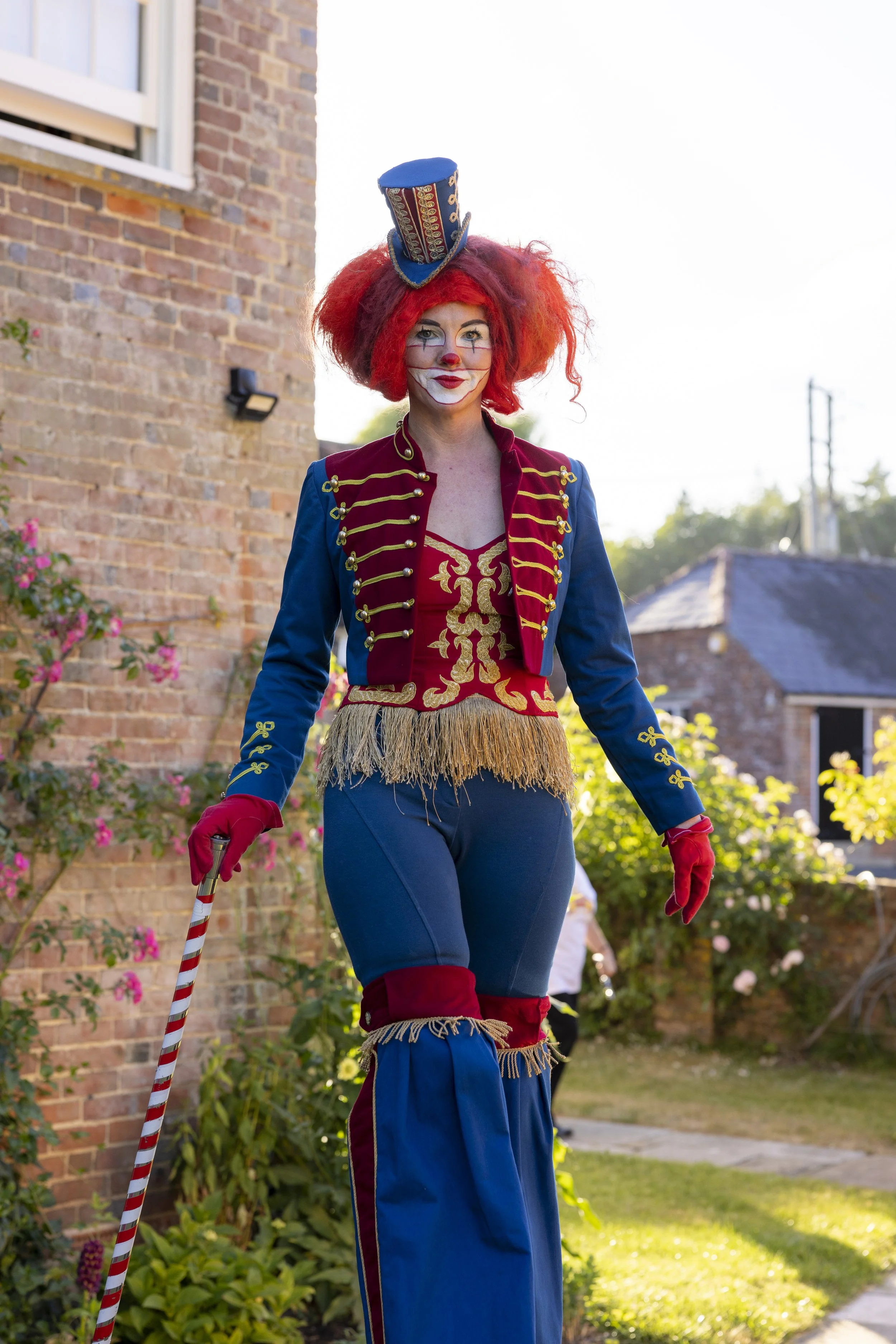 A woman dressed as a clown on stilts, with red curly hair, a blue top hat, red face paint, and a colorful circus costume, standing outdoors in a garden with brick buildings in the background.