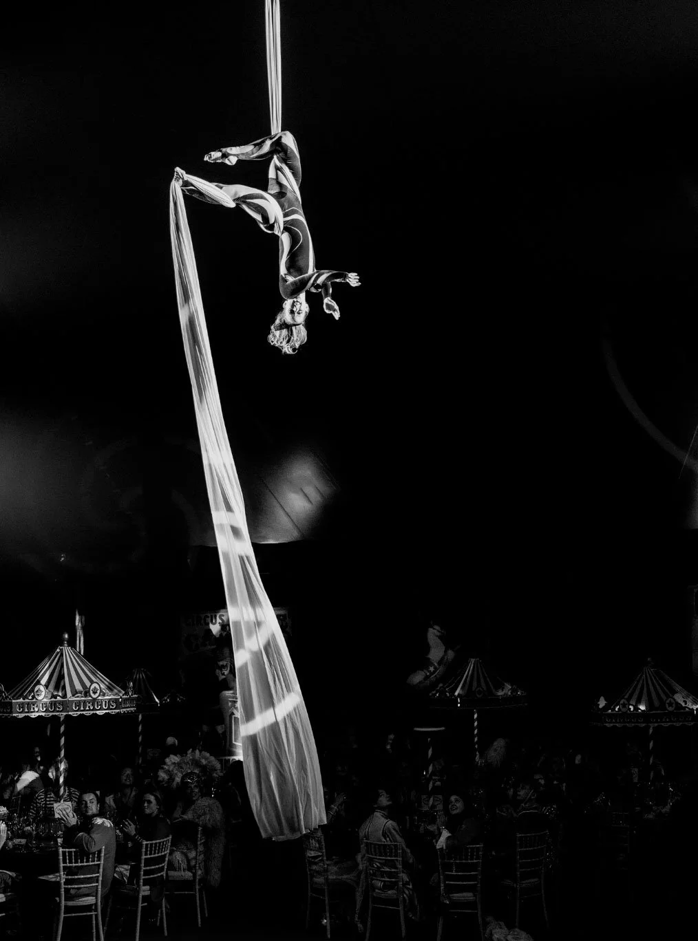 An aerial silks performer in a circus acts upside down on long fabric, hanging from the ceiling of a circus tent, with an audience watching below.