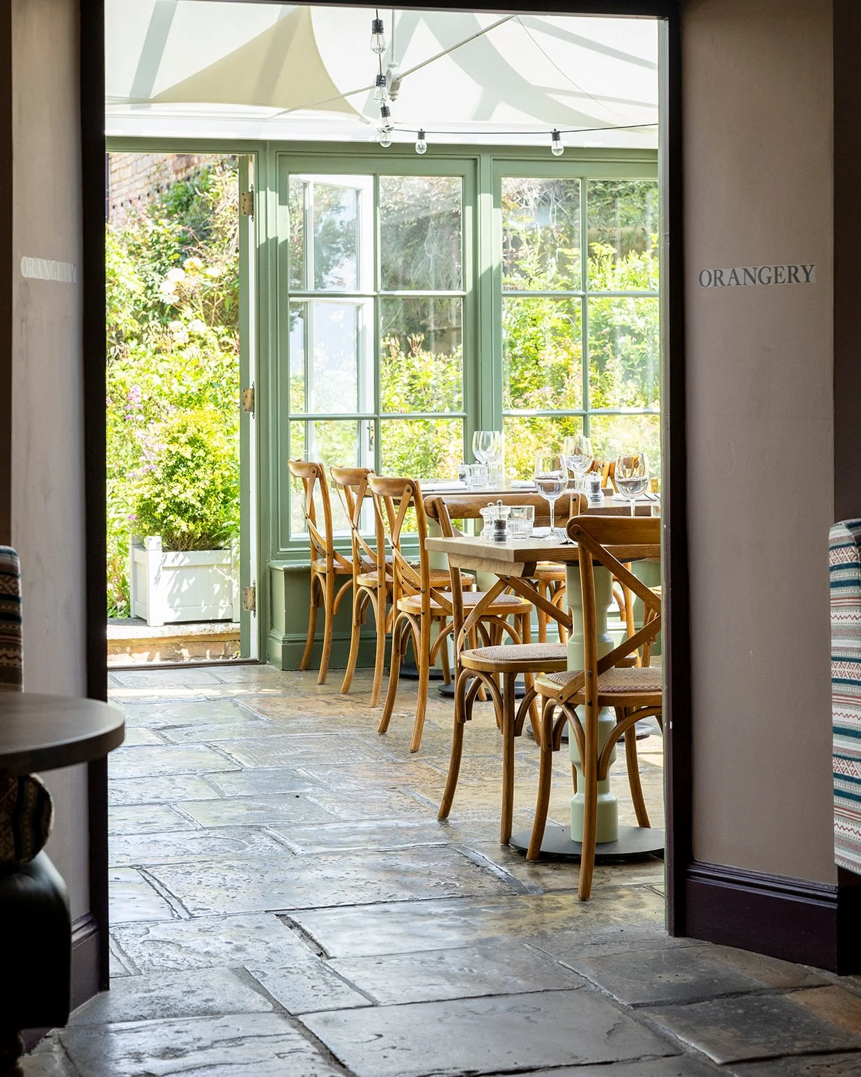 A sunlit dining area with wooden chairs around a table, set with wine glasses and silverware, next to large green-framed windows overlooking a garden.