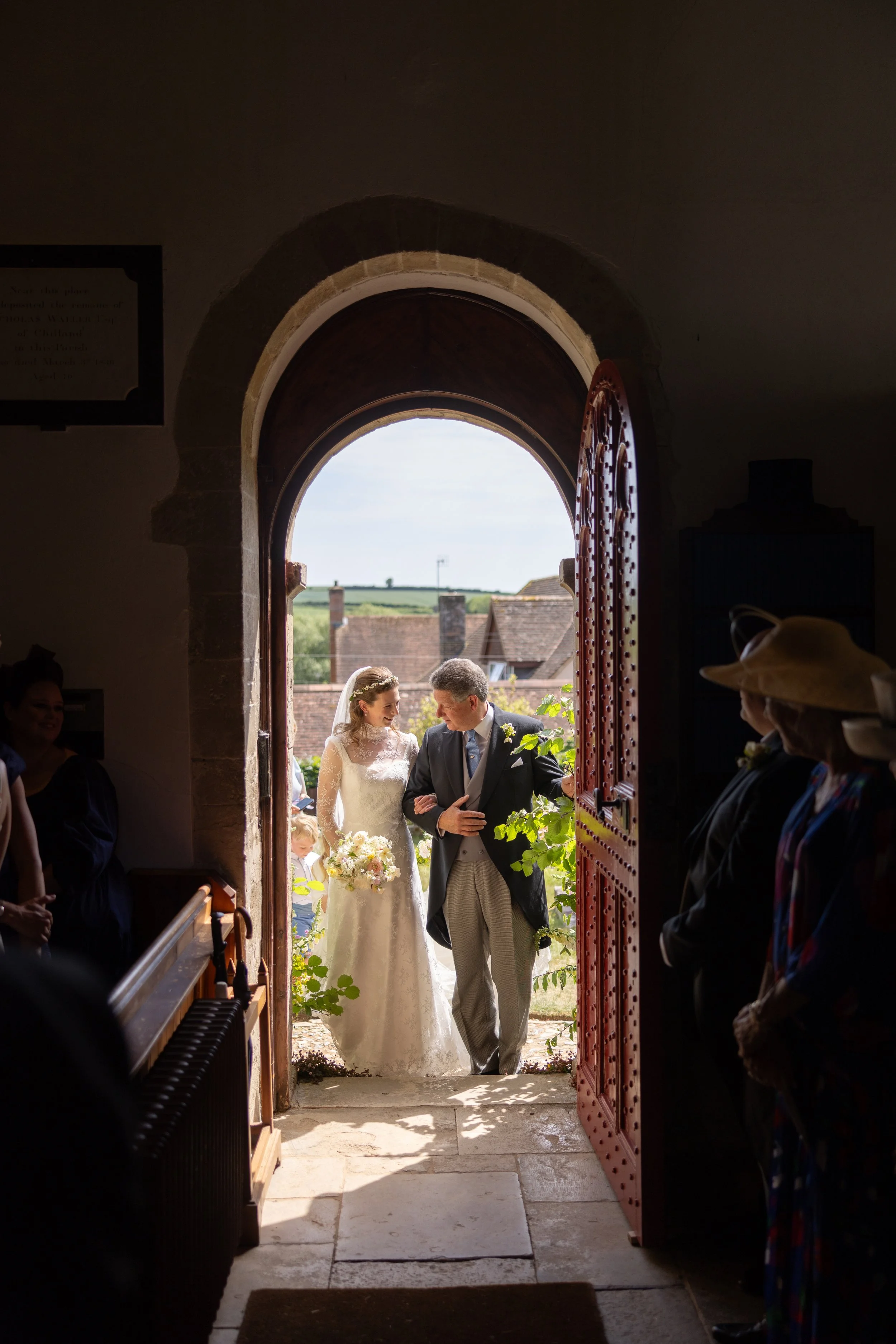 A bride and an older man, likely her father, standing in the doorway of a church, smiling at each other. The bride is holding a bouquet of flowers, and they are about to walk outside. Guests are seated inside, visible on the sides.