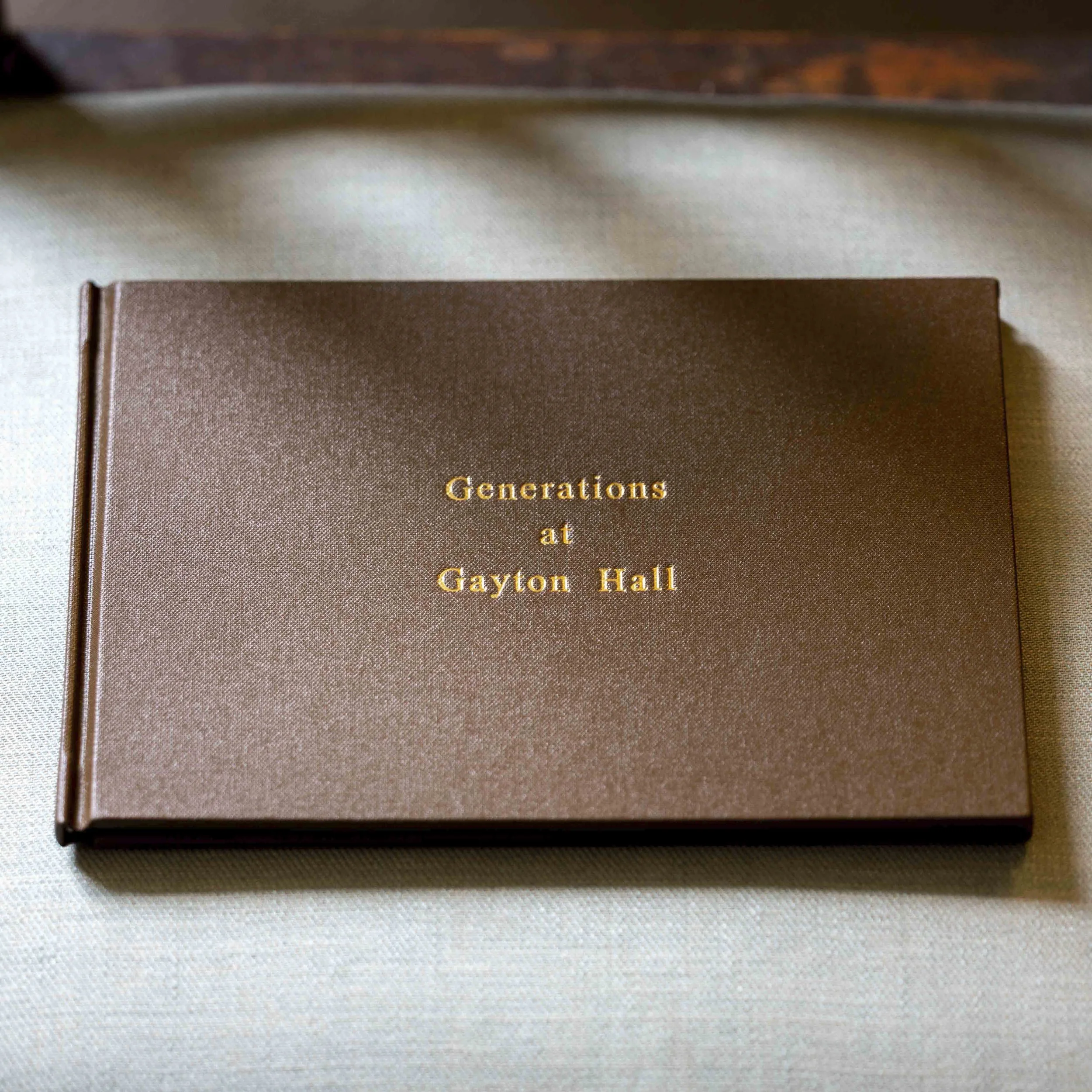 A brown hardcover book titled 'Generations at Gayton Hall' with gold lettering, placed on a light-colored fabric surface.