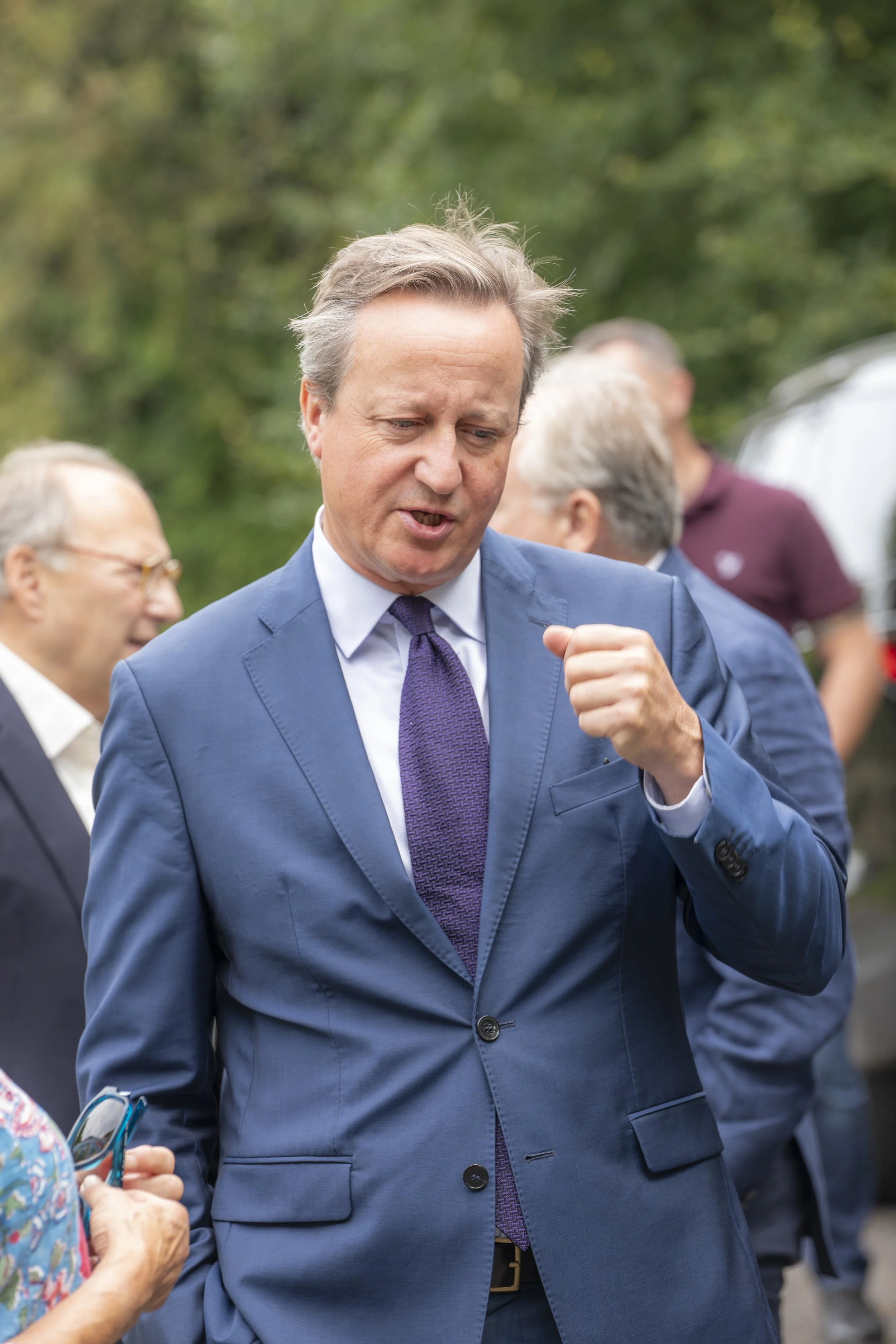 A man in a blue suit and purple tie talking to others outdoors, surrounded by green trees, during daytime.