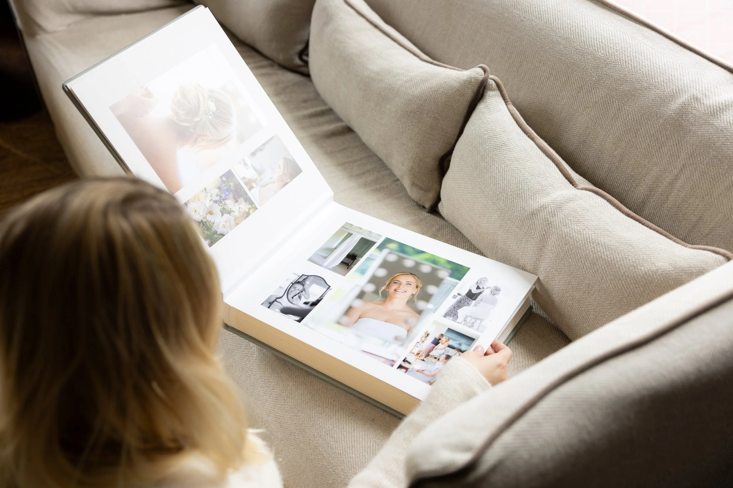 A person with blonde hair looking at a photo album with images of women, flowers, and fashion that is resting on a beige couch.