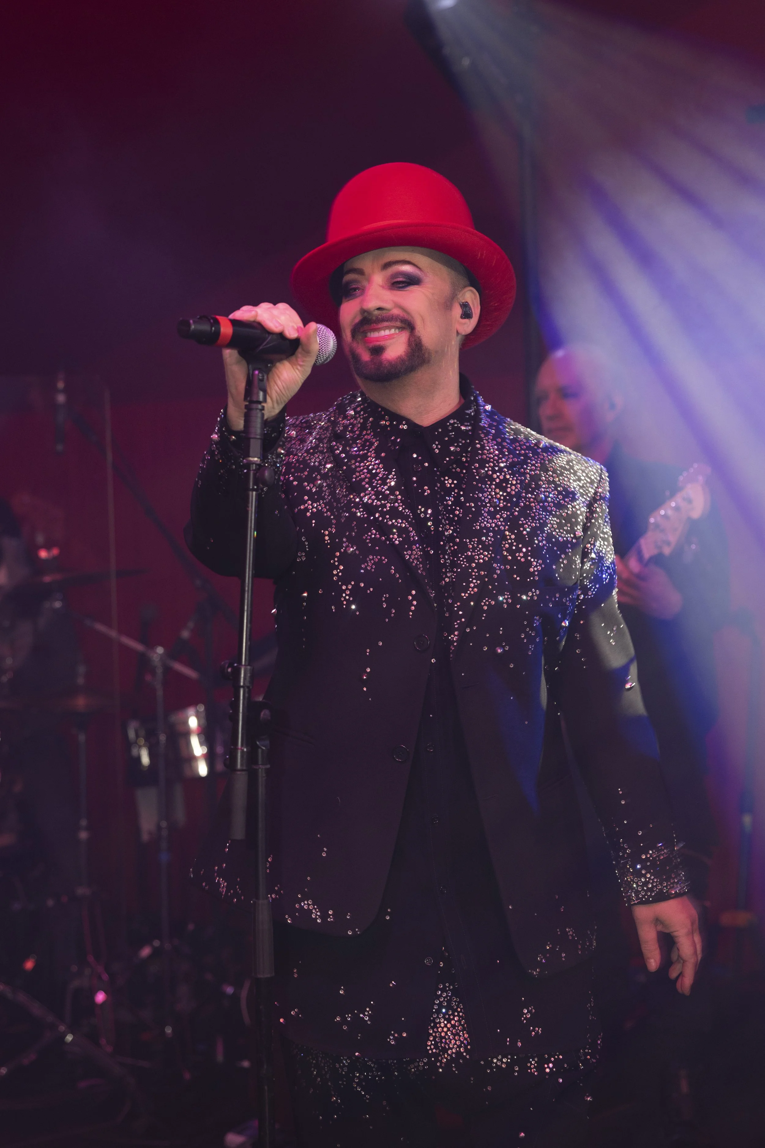A performer on stage wearing a red hat and a black suit adorned with sparkling embellishments, singing into a microphone.