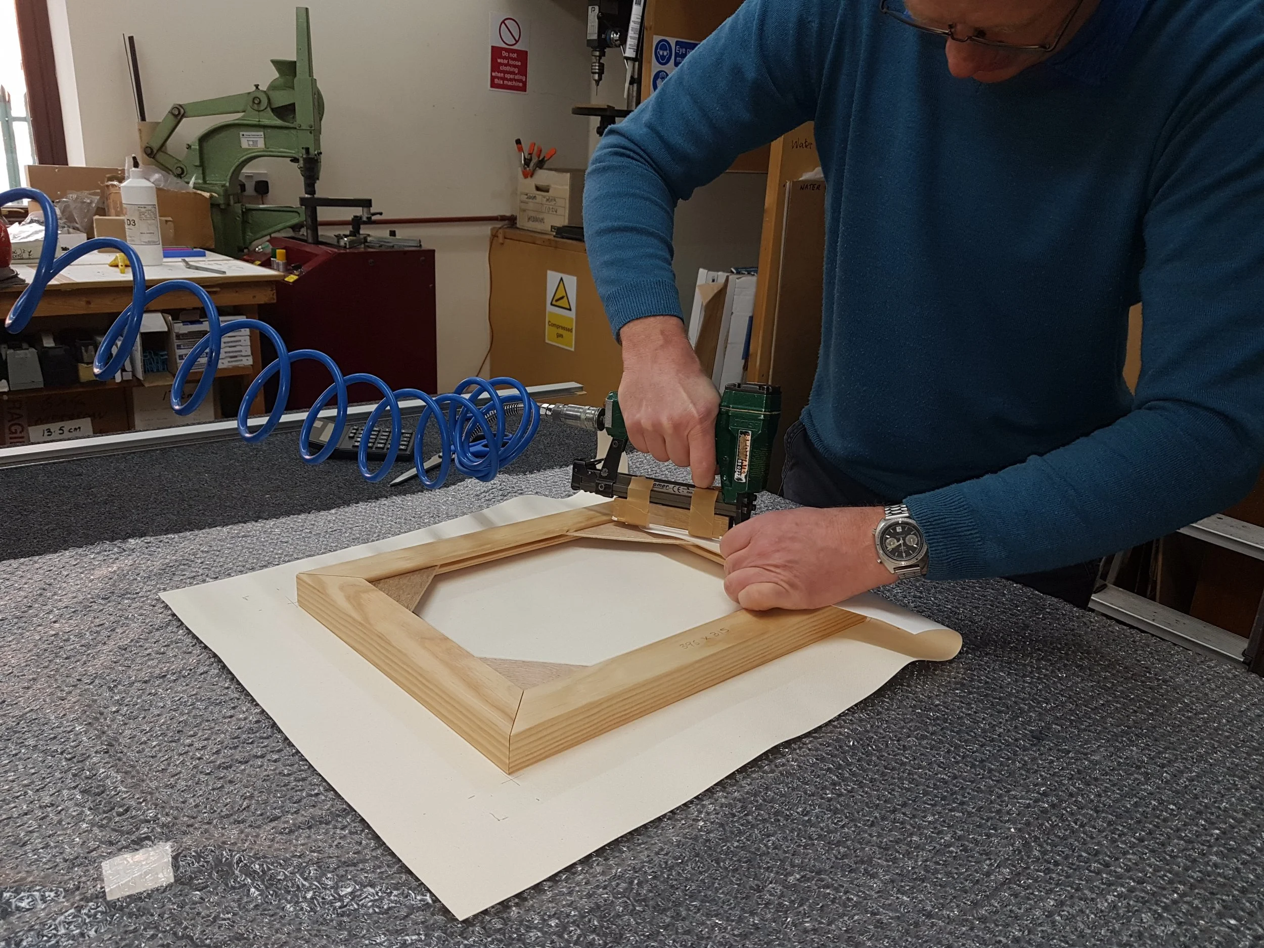 A person in a blue sweater working in a workshop, assembling a wooden picture frame using a power tool, with workshop tools and equipment visible in the background.
