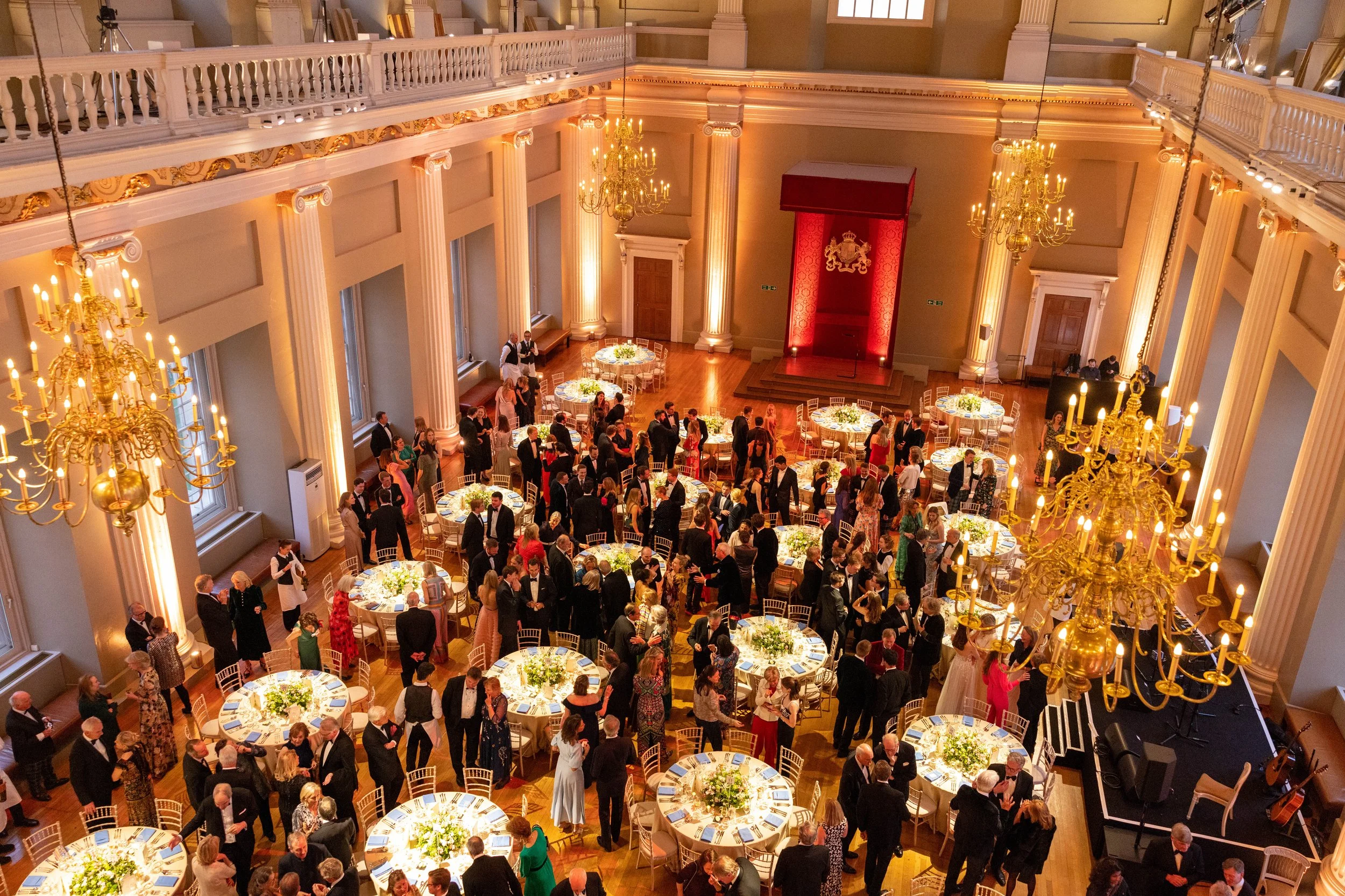 Elegant ballroom with chandeliers, tall columns, and a stage with a curtain featuring a crown emblem. Guests in formal attire are mingling and dining at round tables decorated with floral centerpieces.