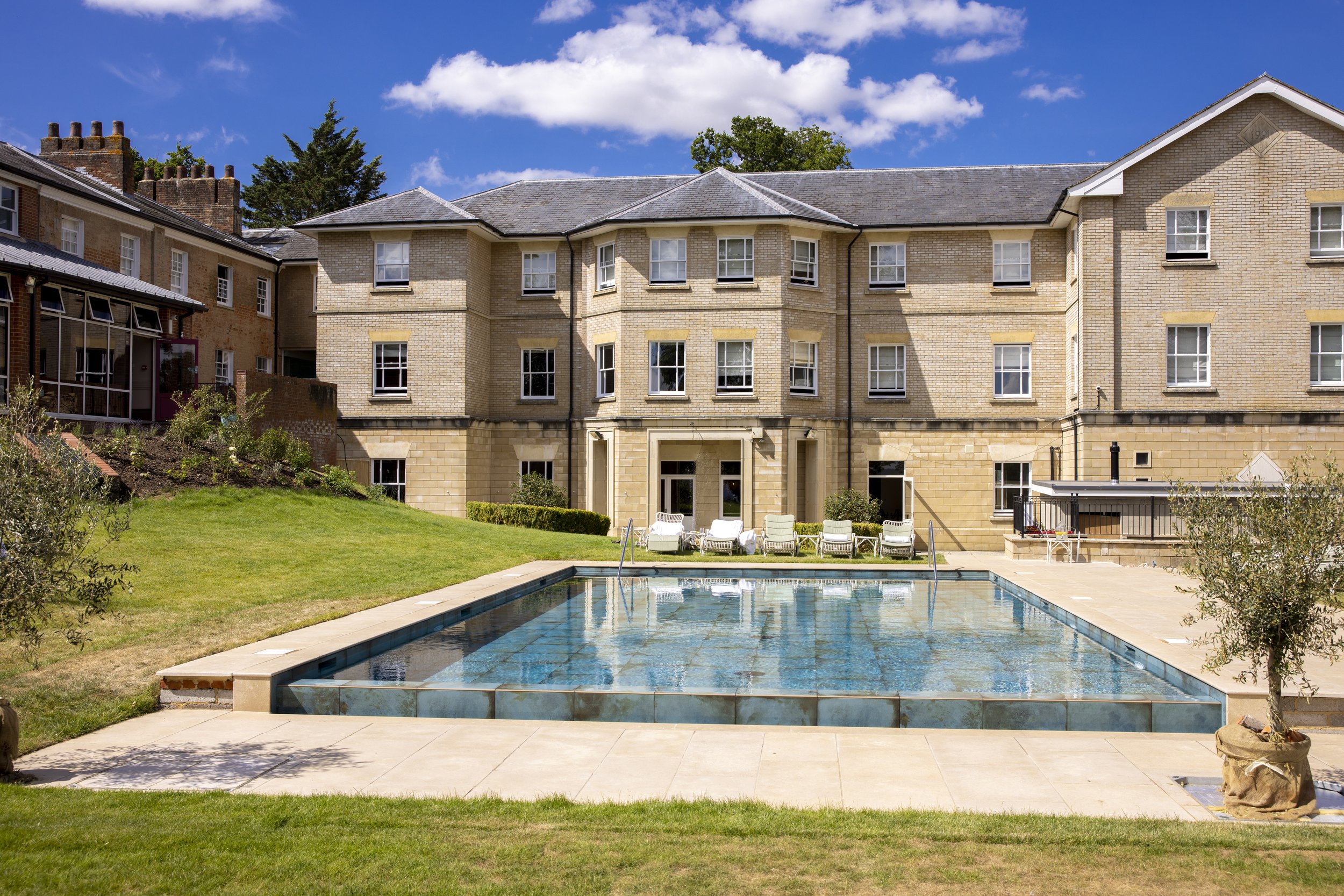 A residential apartment complex with a courtyard and swimming pool, surrounded by green grass and trees, under a partly cloudy blue sky.