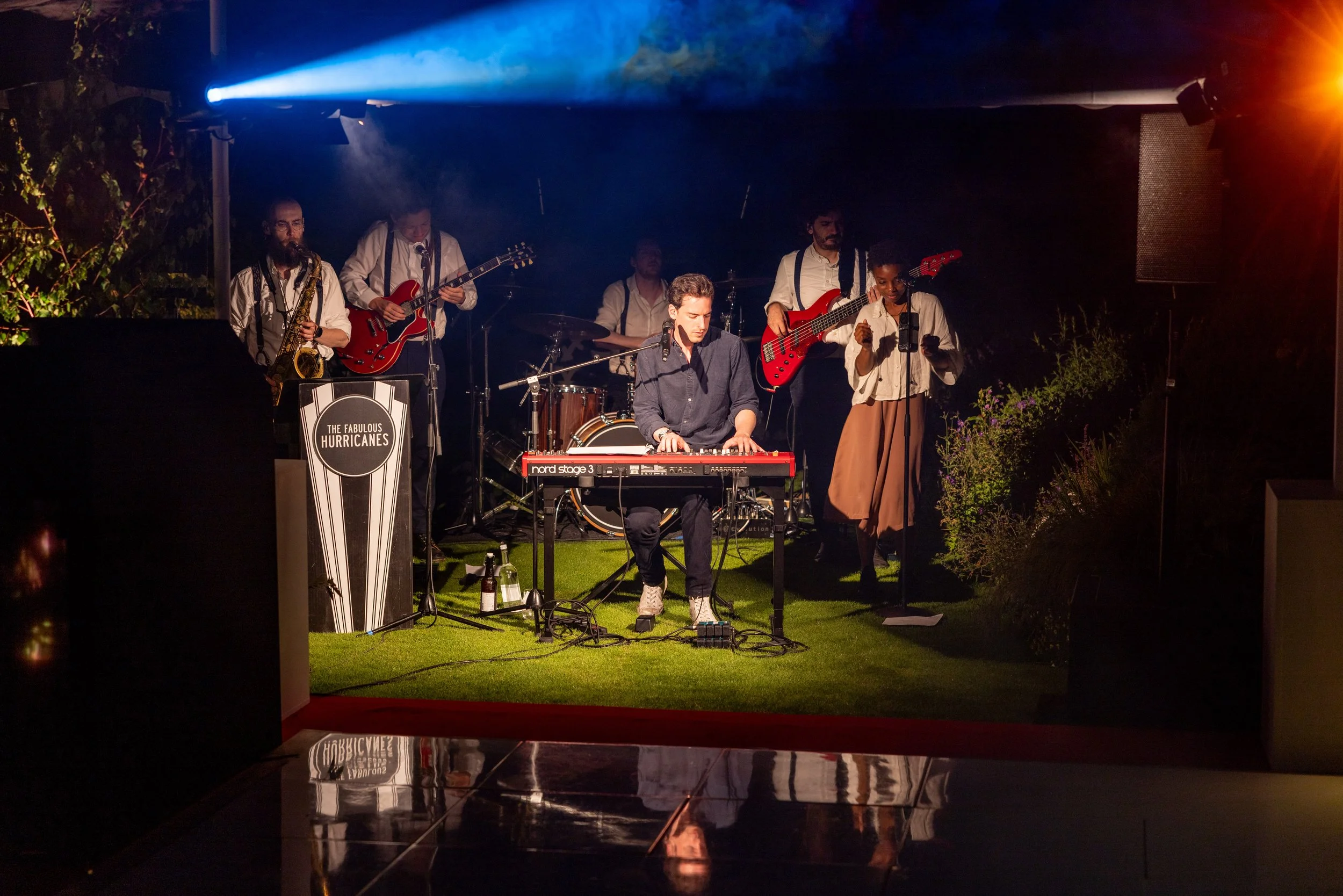 A band performs on a small outdoor stage at night, with a keyboard player in the center, guitarists and a singer, and a sign that reads 'The Fabulous Hurricanes'.