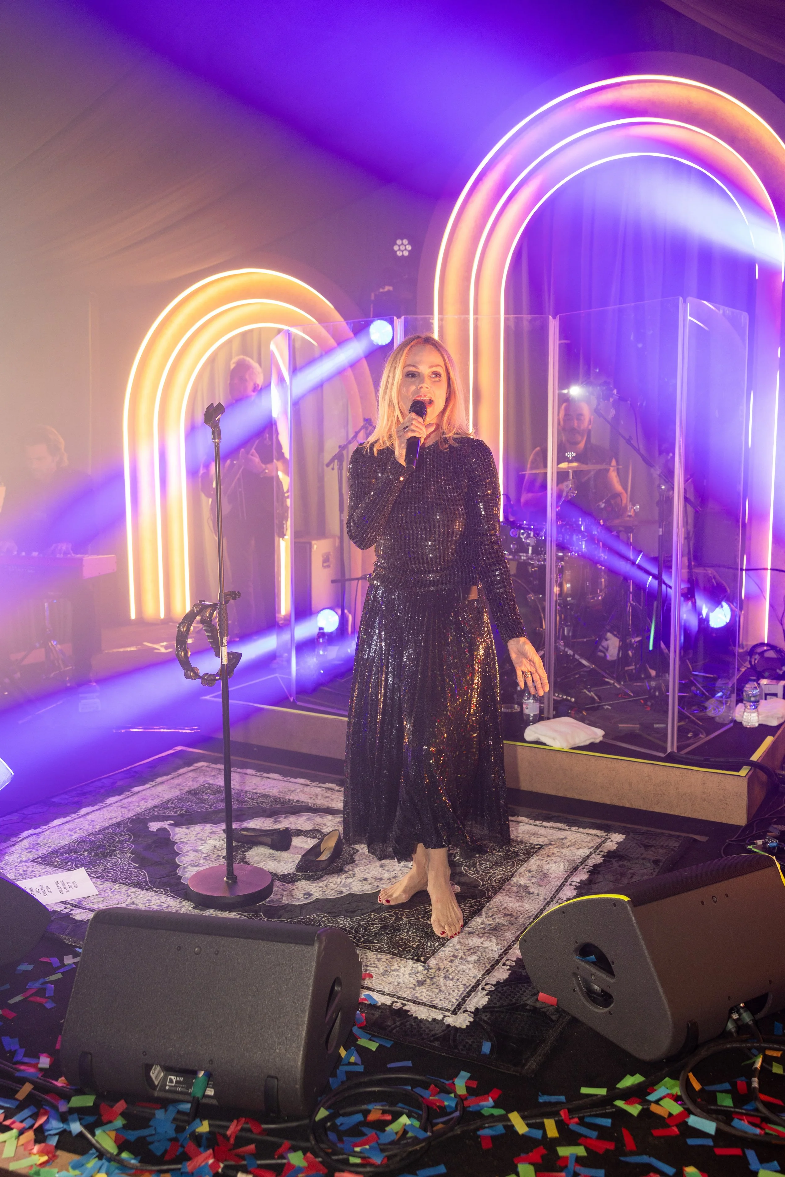 A woman in a black sequined dress singing into a microphone on stage with colorful neon rainbow arches in the background, band members, and confetti on the floor.