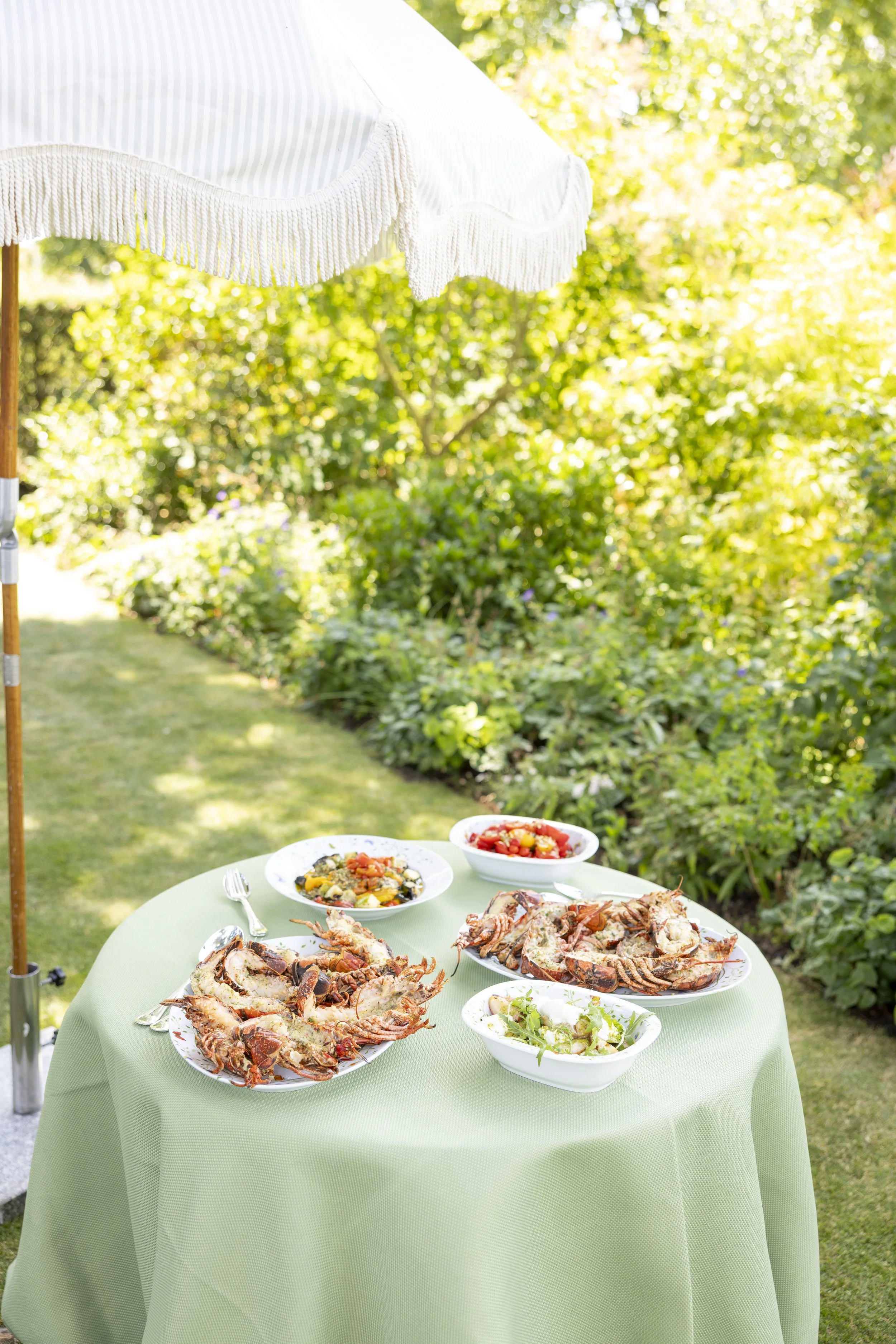 Outdoor table with cooked lobsters, salad, and bowls of side dishes on a green tablecloth, set in a garden with sunlight and greenery in the background.