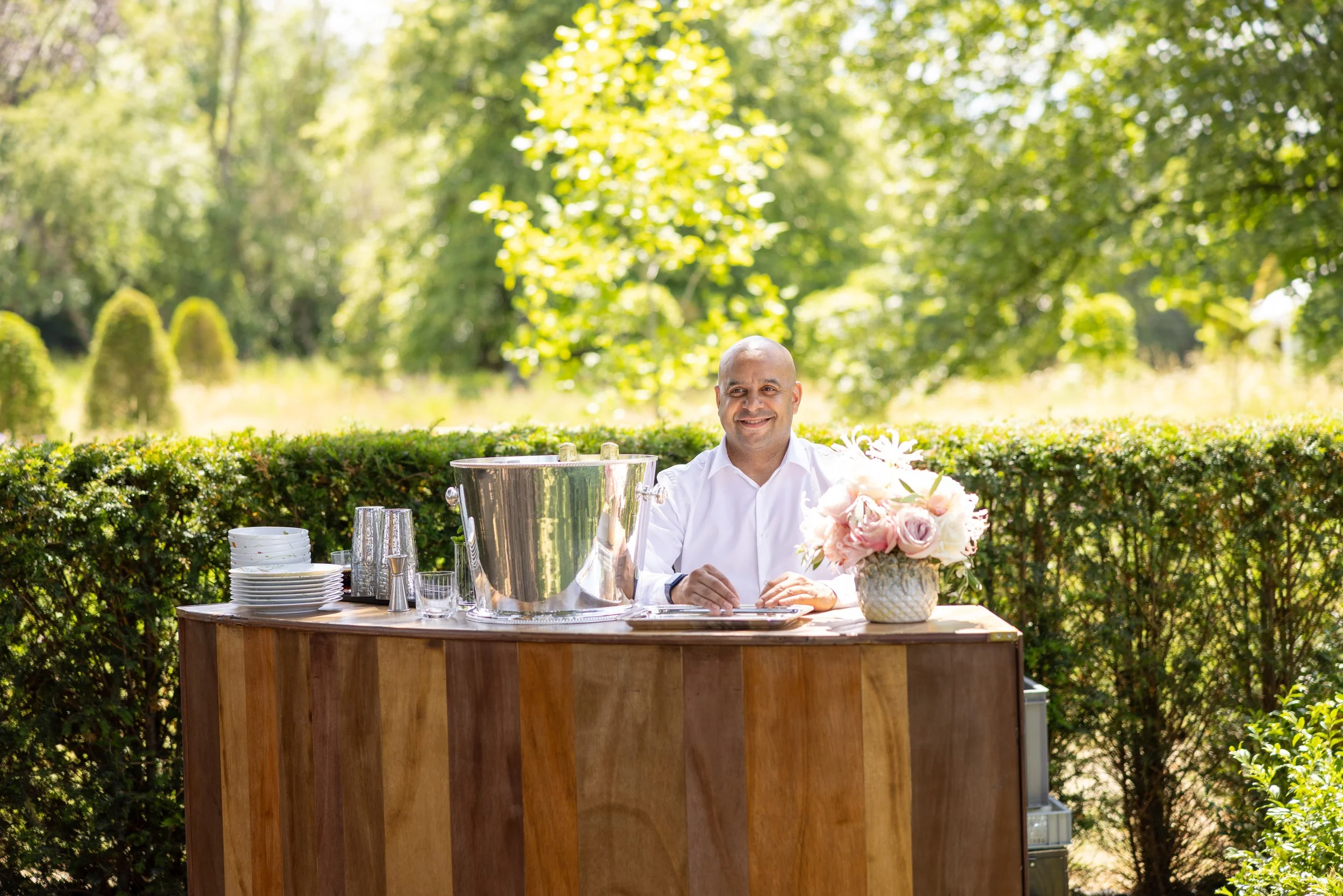 A smiling man in a white shirt seated behind an outdoor bar with a large metal ice bucket, glassware, and a vase of pink roses, set against a lush green background of trees and bushes.