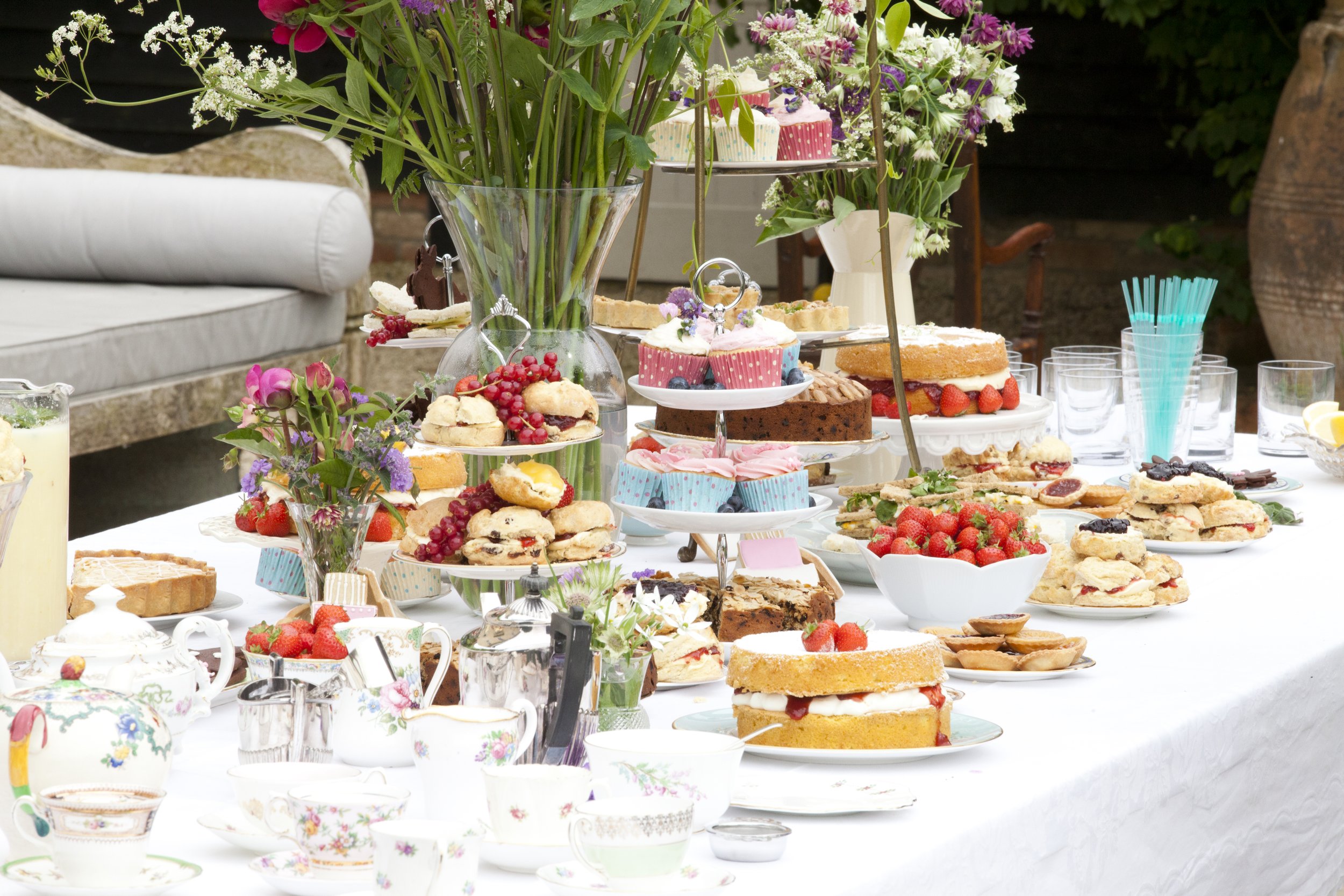 A table set with various desserts, including cakes, cookies, strawberries, and cupcakes, along with flowers and cups, at an outdoor gathering.