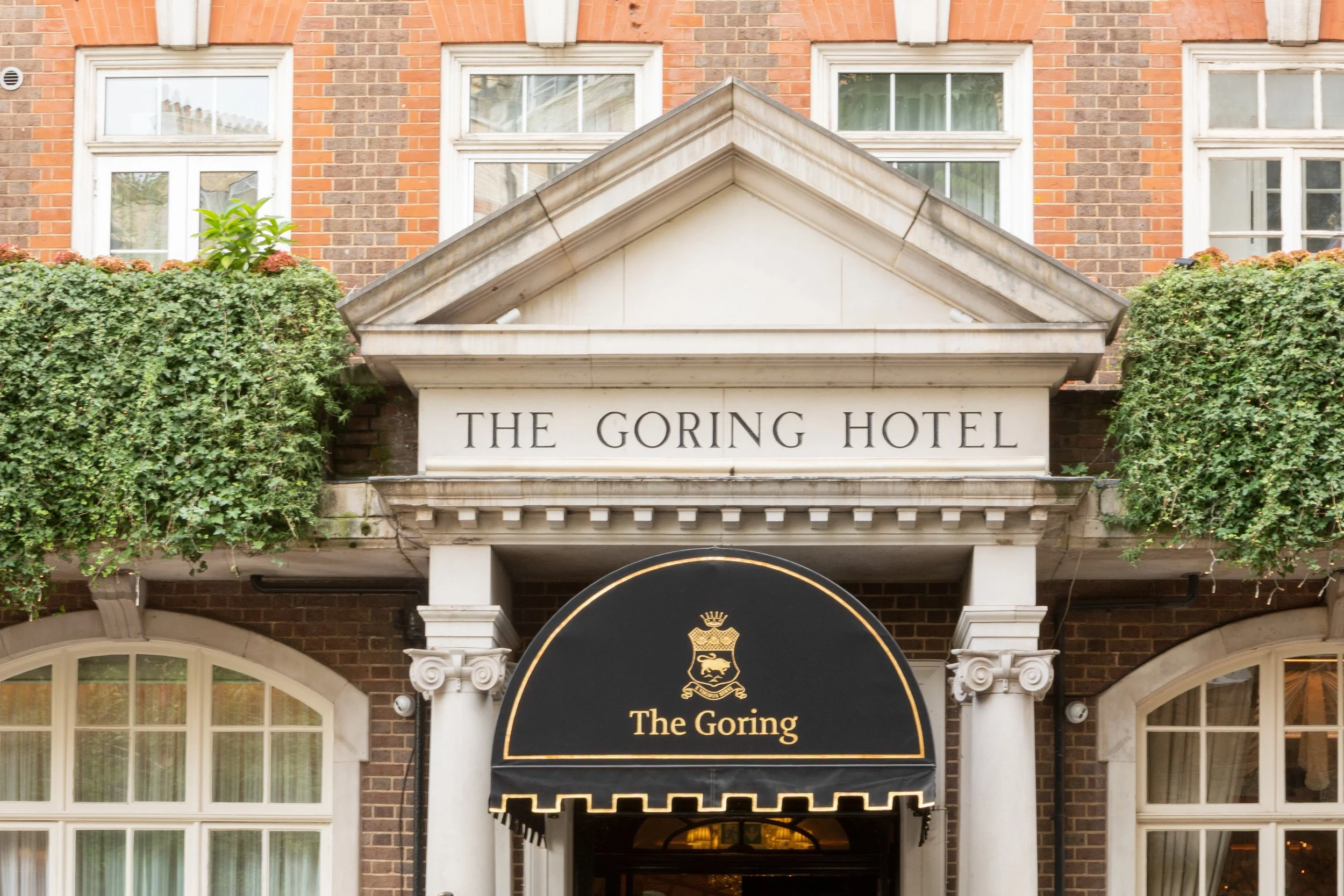 The entrance of The Goring Hotel with a black awning featuring gold lettering and crest, and a brick building with white-framed windows.