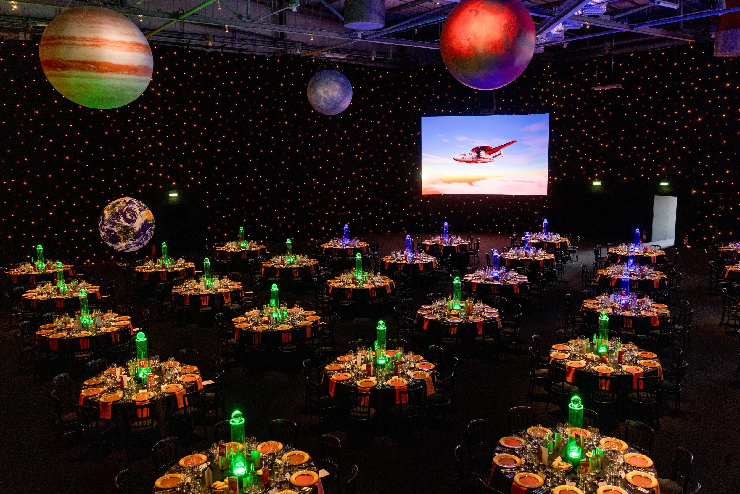 A large event hall decorated with planets hanging from the ceiling and world-themed lighting, with tables set for a banquet.