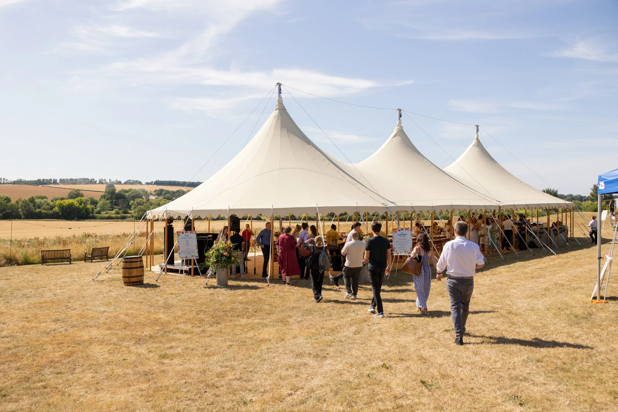 People gathering under a large white event tent in an open field on a sunny day.