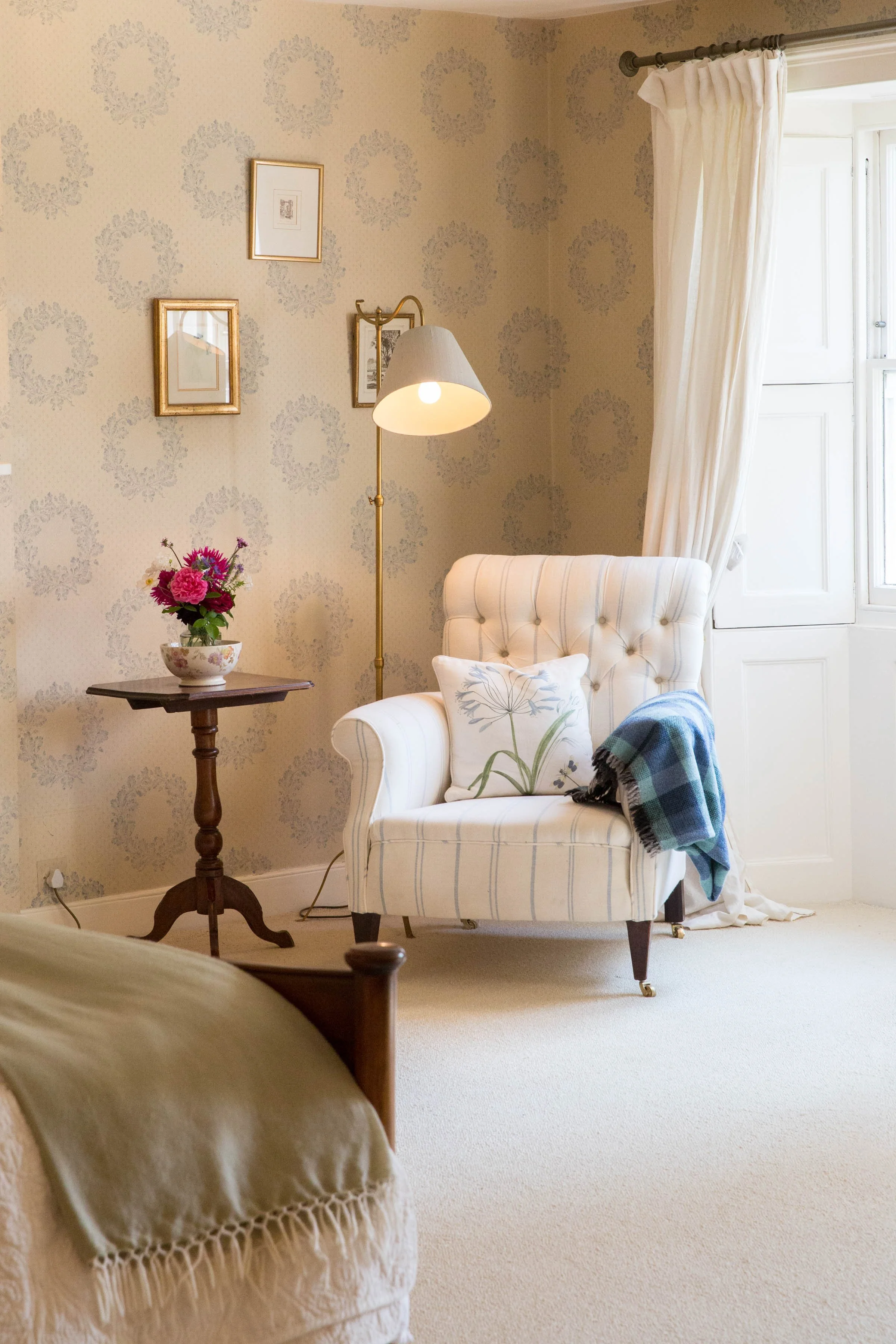 A cozy corner of a living room with a white upholstered armchair, a decorative pillow, a plaid blanket, a small wooden side table with a flower vase, a standing lamp, and framed pictures on the wall near a window with white curtains.