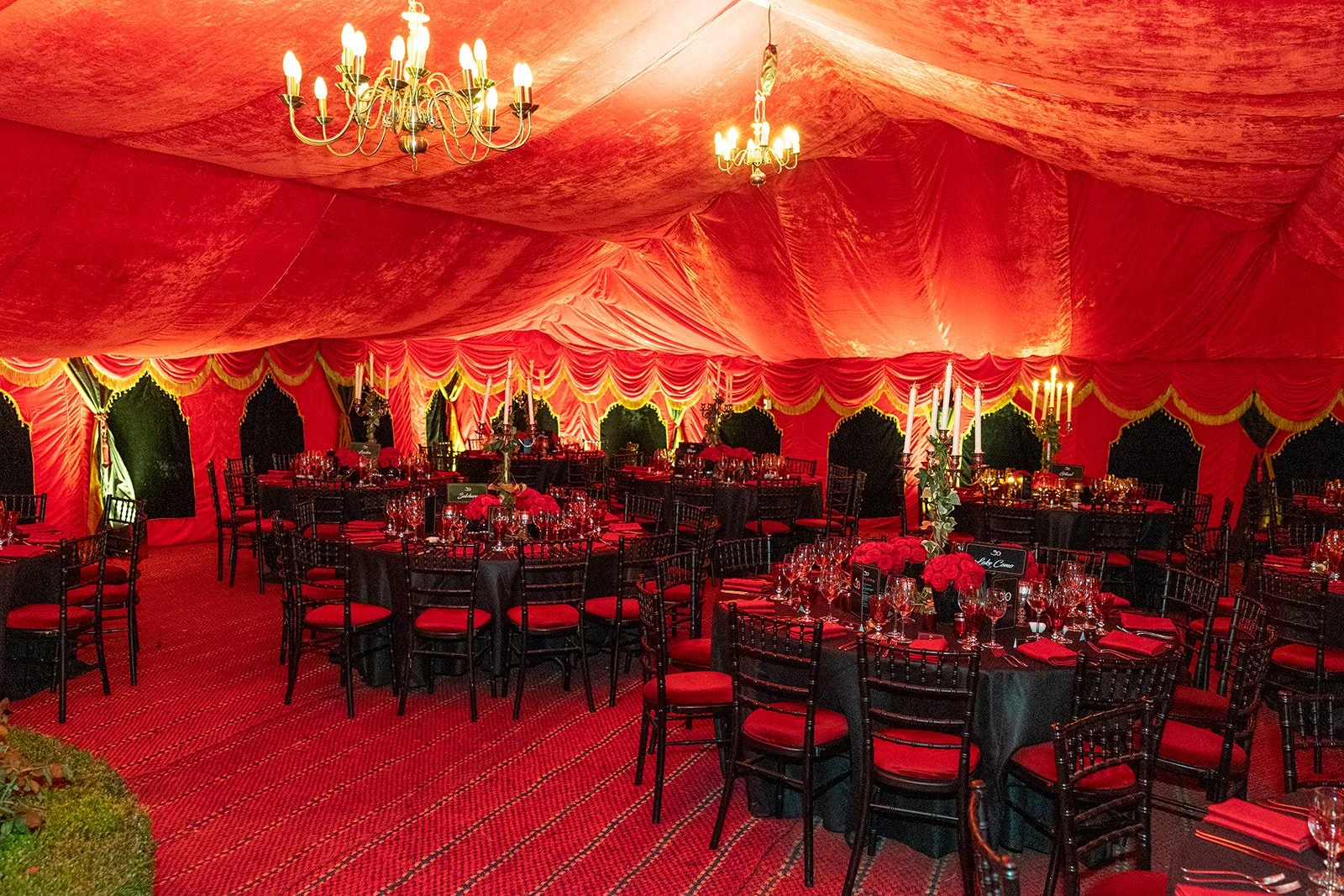 Elegant event tent with red drapery, chandeliers, and round tables decorated with red flowers and candles, set for a celebration or formal gathering.