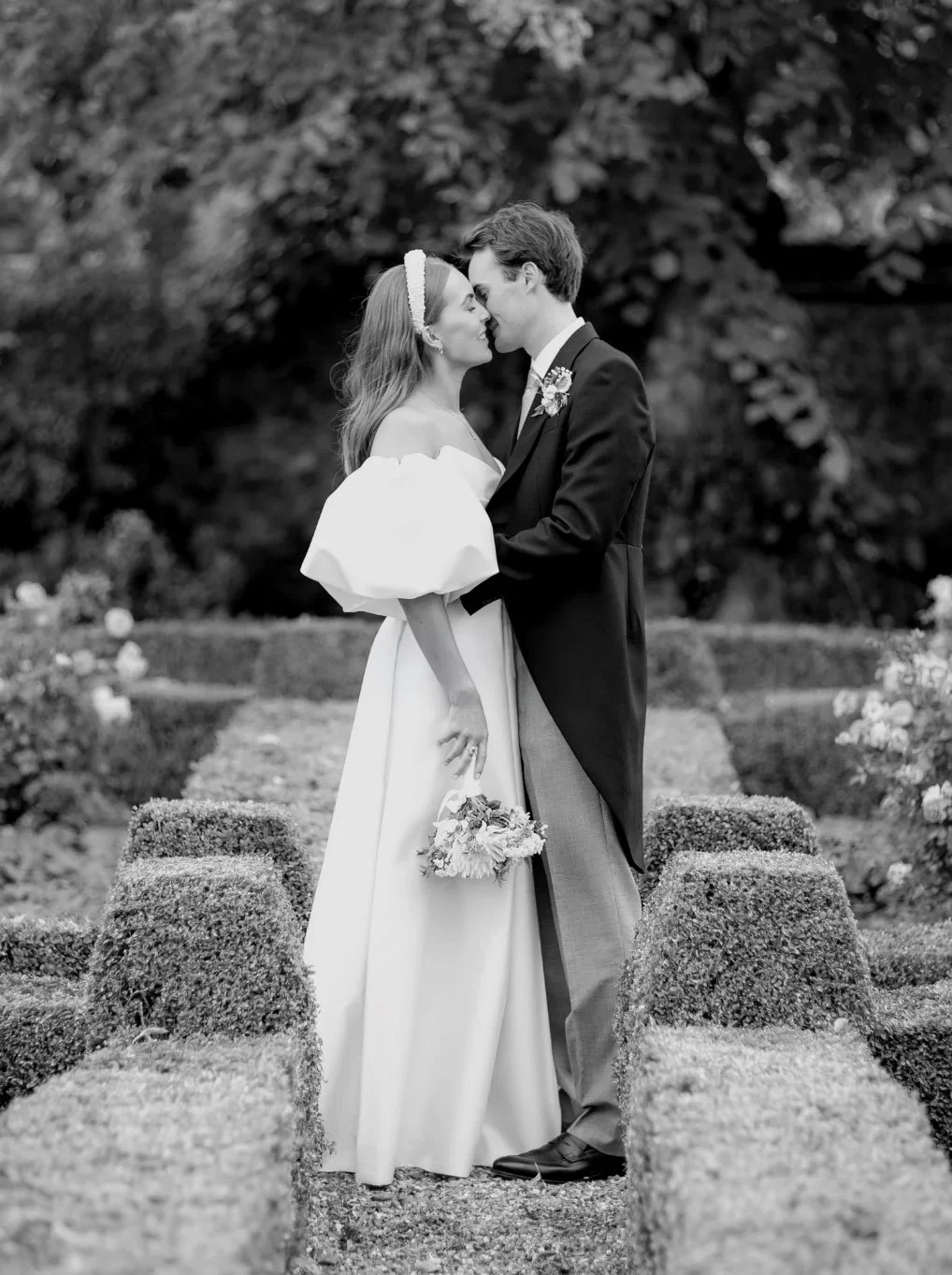 A black and white photo of a bride and groom standing close together in a garden. They are touching foreheads and holding hands, with the bride holding a small bouquet. The garden has topiary and flowers, with trees in the background.