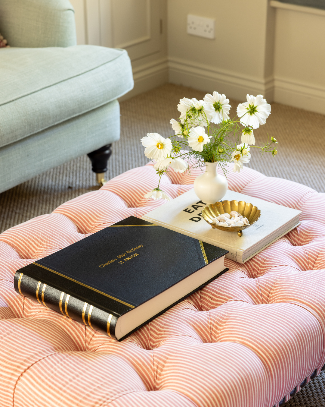 A pink tufted ottoman with a black and gold wedding album, a white book with black text, a small gold dish with white candies, and a white vase with white flowers on top, in a living room setting.