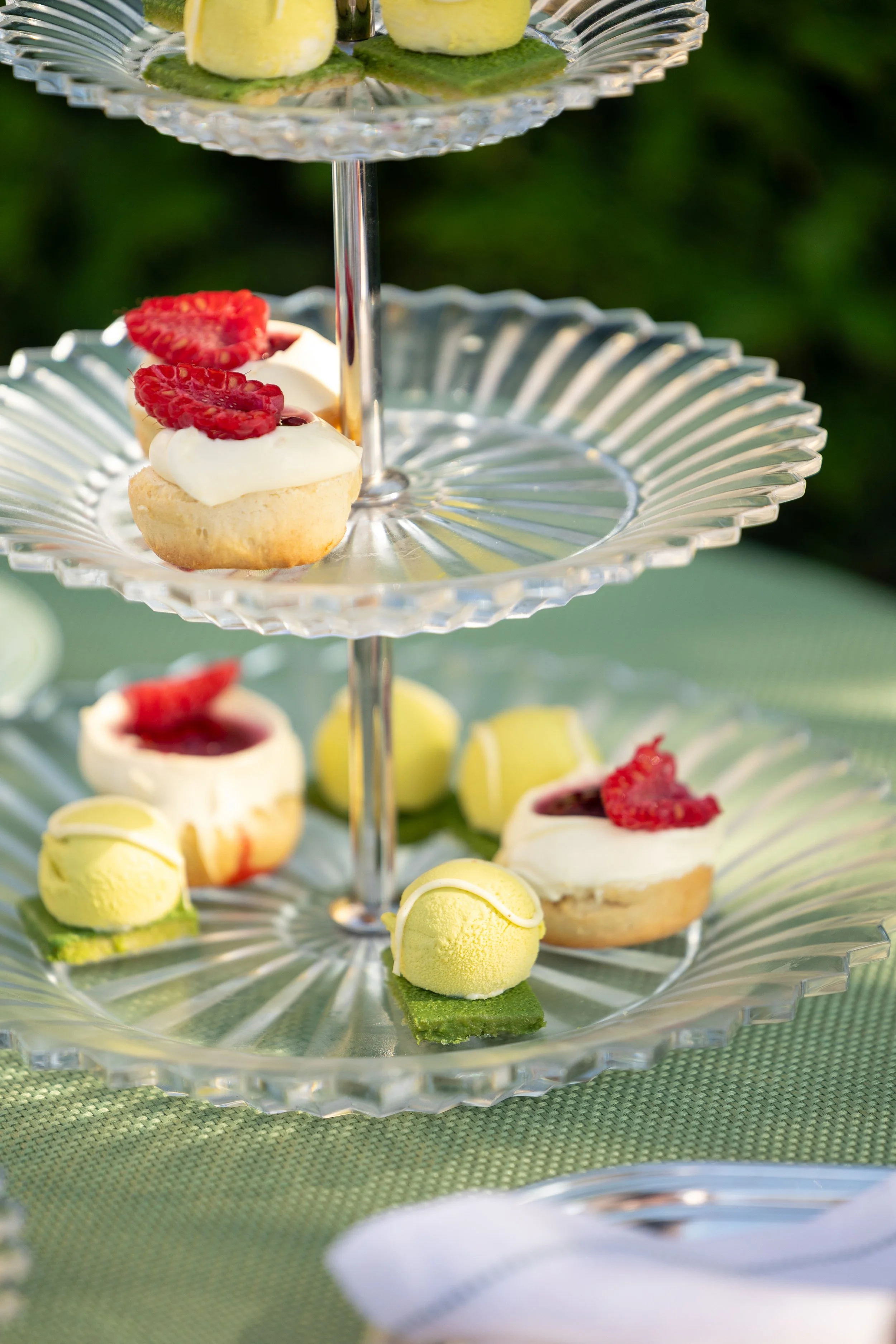 Three-tiered glass dessert stand with assorted pastries and sweets, including small cakes topped with strawberries and raspberries, and yellow macarons on a green tablecloth.