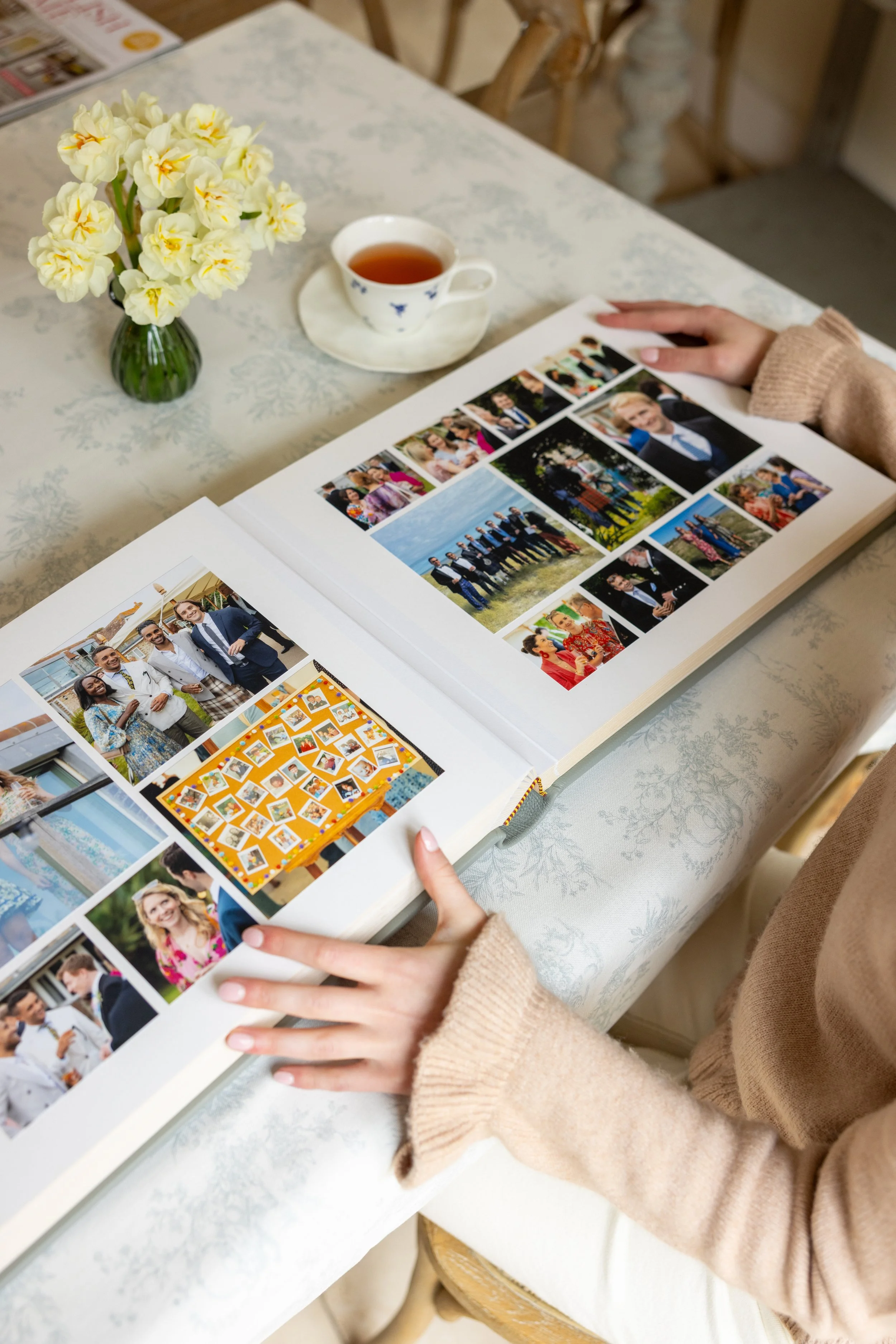 A person looking at a photo album on a table with a vase of yellow flowers and a cup of tea.