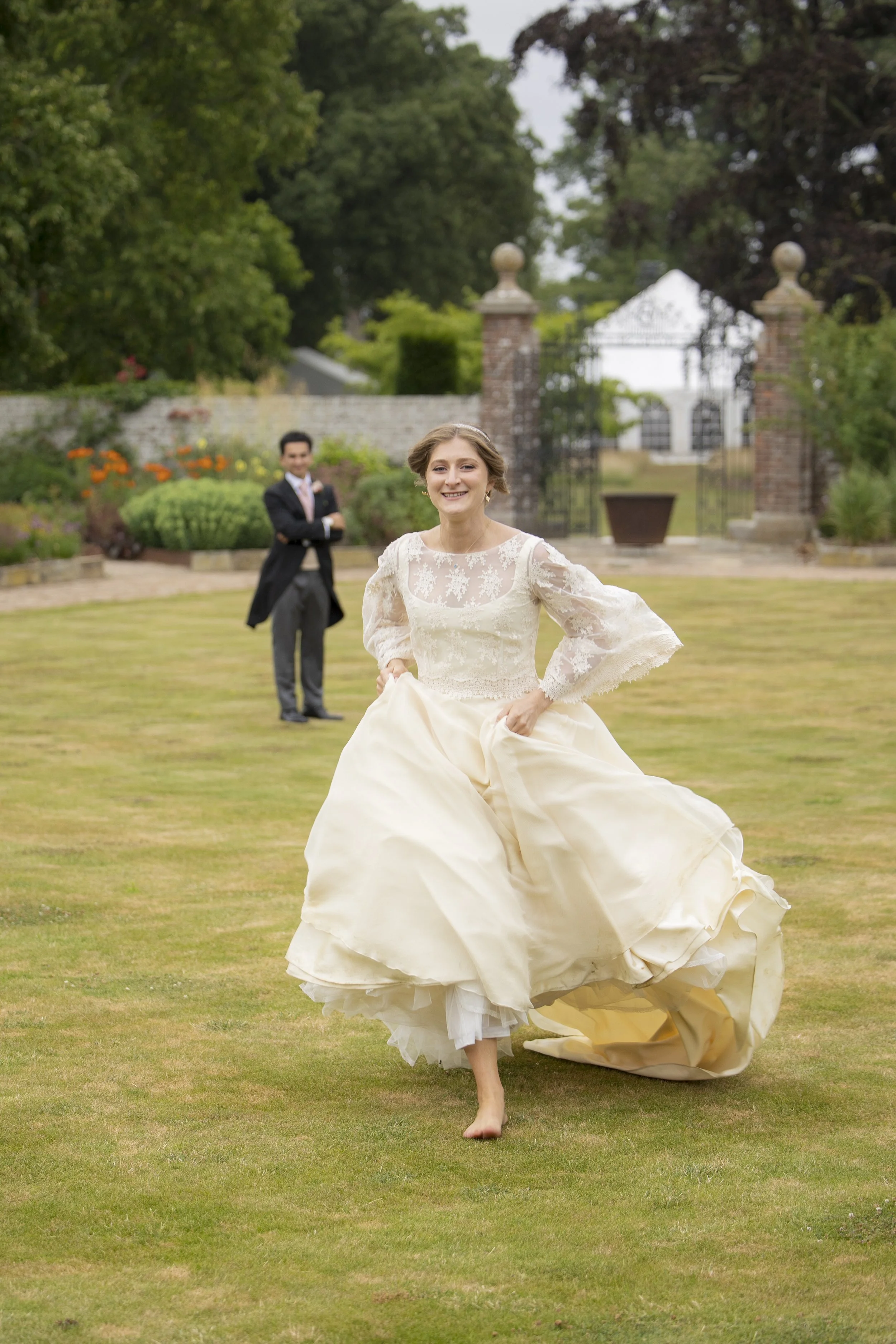 A bride in a wedding dress running on a lawn with a man in a tuxedo standing in the background, at an outdoor wedding venue with trees and a decorative gate.
