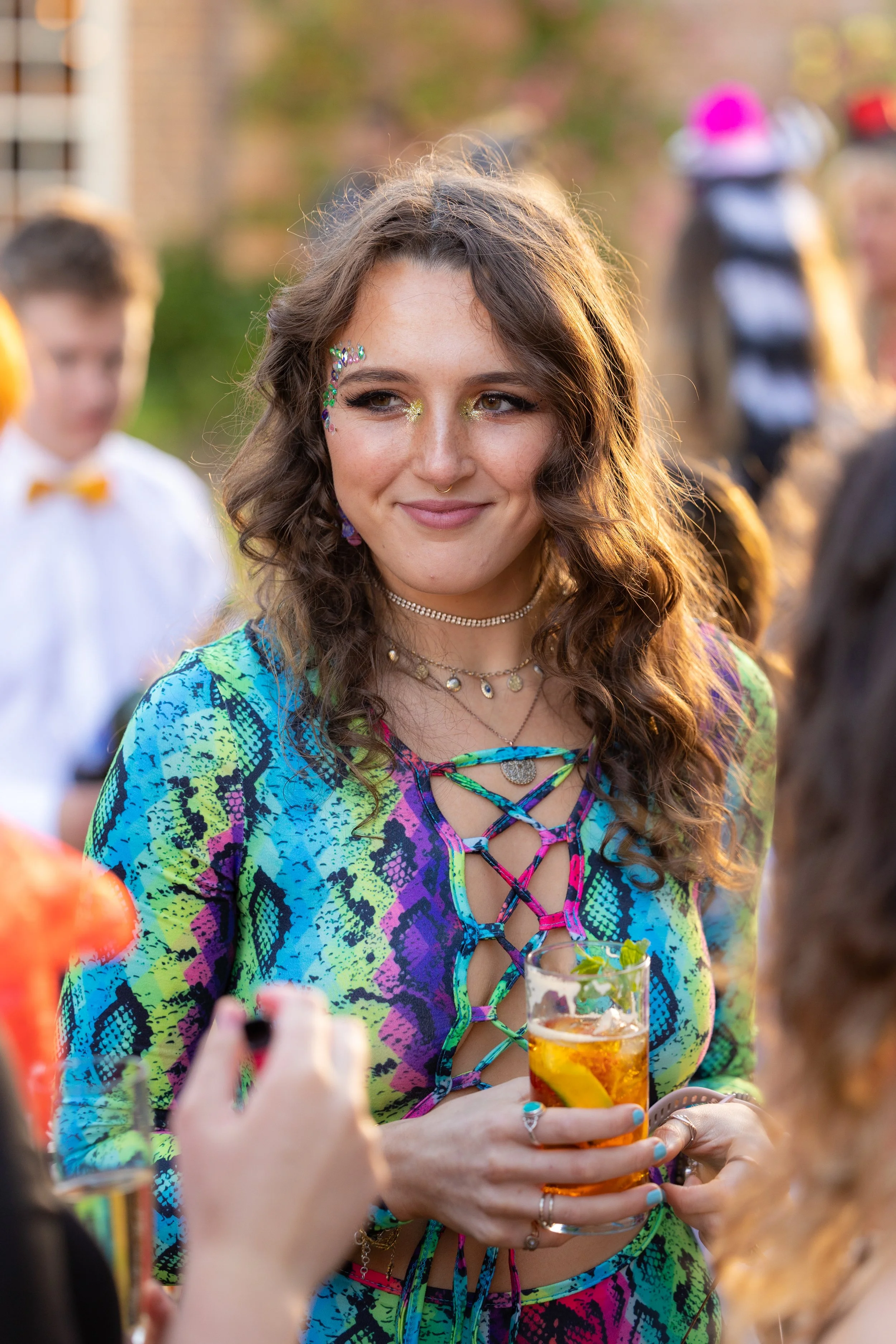 Young woman with colorful, patterned lace-up top, holding a drink, at an outdoor event with other people