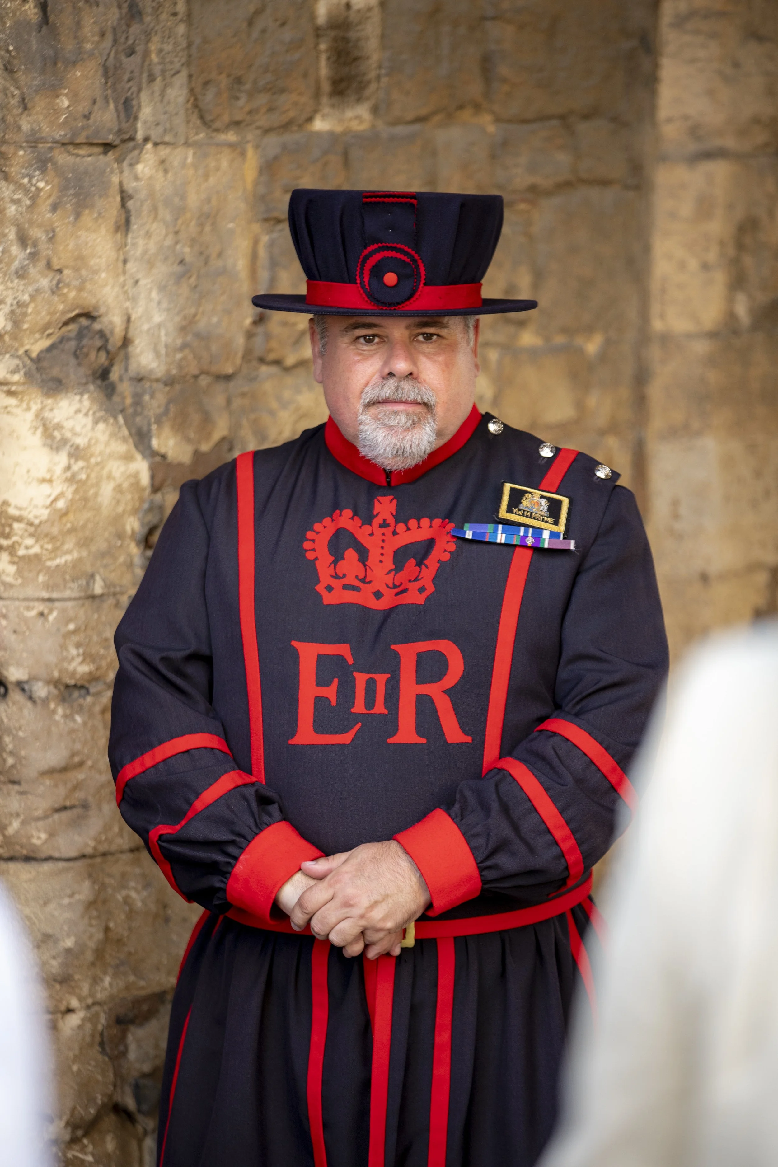 A man dressed in a historical uniform resembling the British Royal Engineers, wearing a black and red outfit with a tall black hat and a badge, standing against a stone wall.