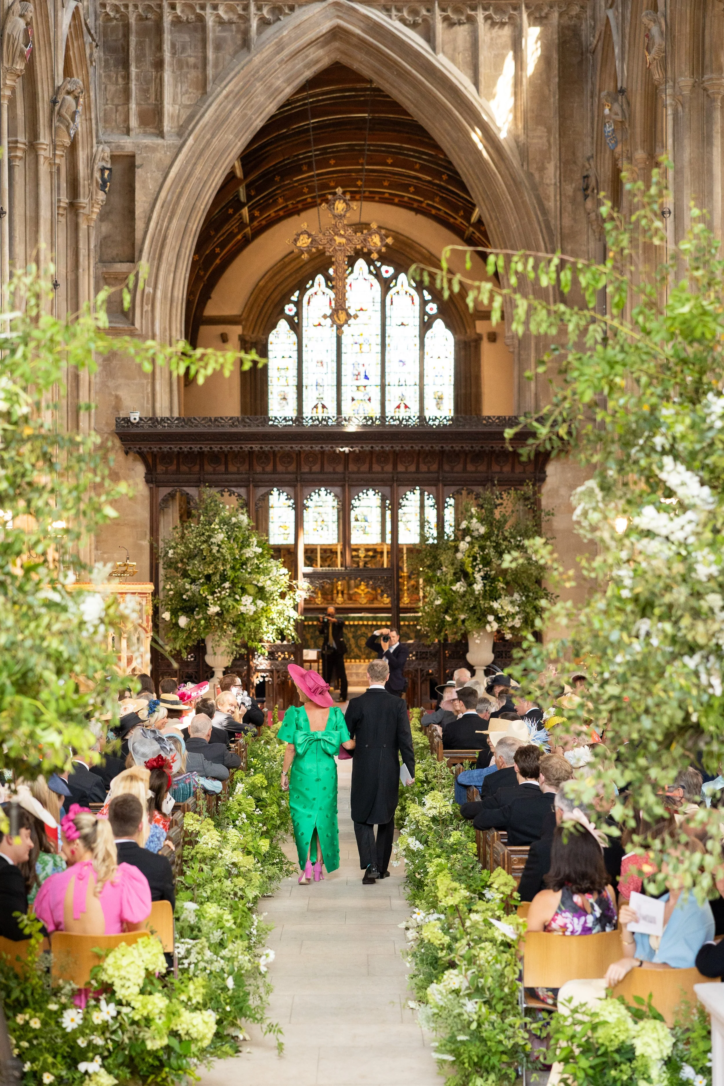 Inside a historic church during a wedding ceremony, guests seated on wooden benches, a couple walking down the aisle, decorated with green foliage and white flowers, with stained glass windows and a large chandelier hanging from the arched ceiling.