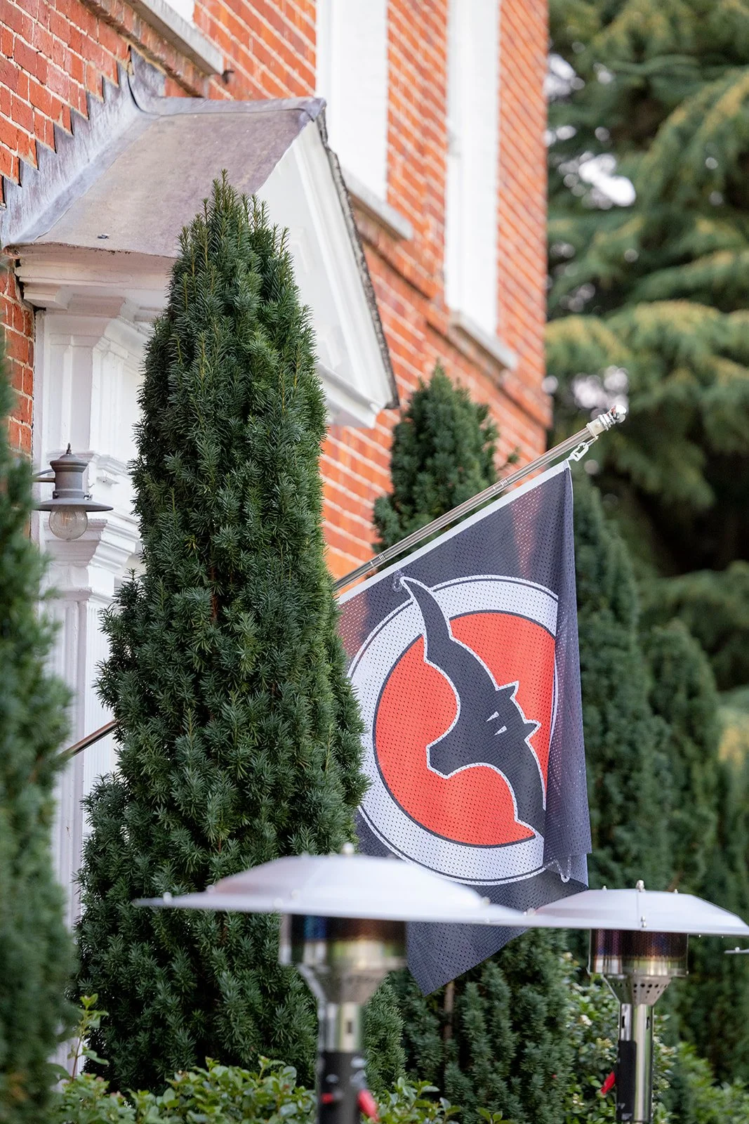 Exterior of a brick house with white trim, decorated with tall evergreen trees and a flag featuring a unicorn silhouette on a red background. Patio heaters are visible in the foreground.