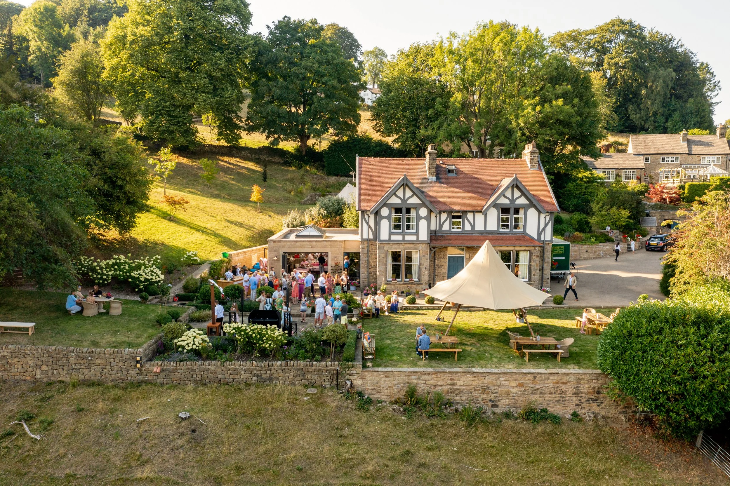 An outdoor gathering on the lawn of a large house with a Tudor-style facade, featuring people socializing, a white canopy tent, and outdoor seating on a sunny day with green trees and a hillside in the background.