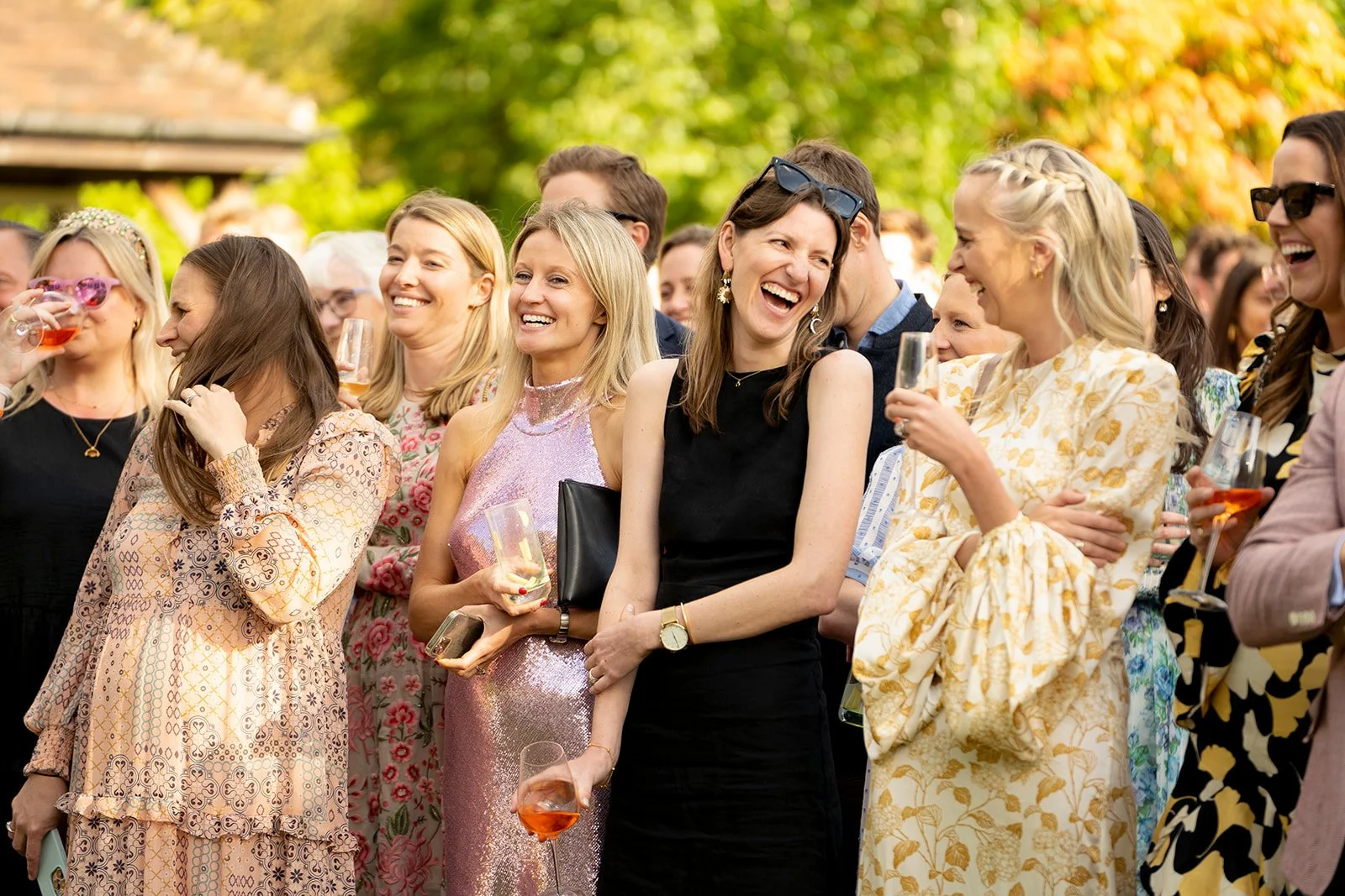 A group of women gathered outdoors, smiling and laughing while holding glasses of champagne or wine at a celebration or social event.