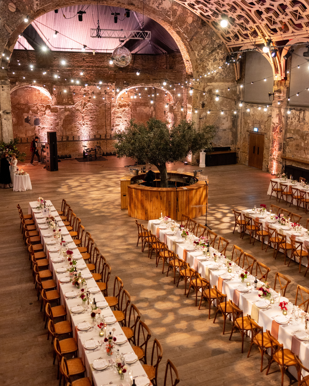 Long banquet tables with white tablecloths and floral centerpieces in a rustic event hall with exposed brick walls, string lights, and a large central tree in a wooden planter.